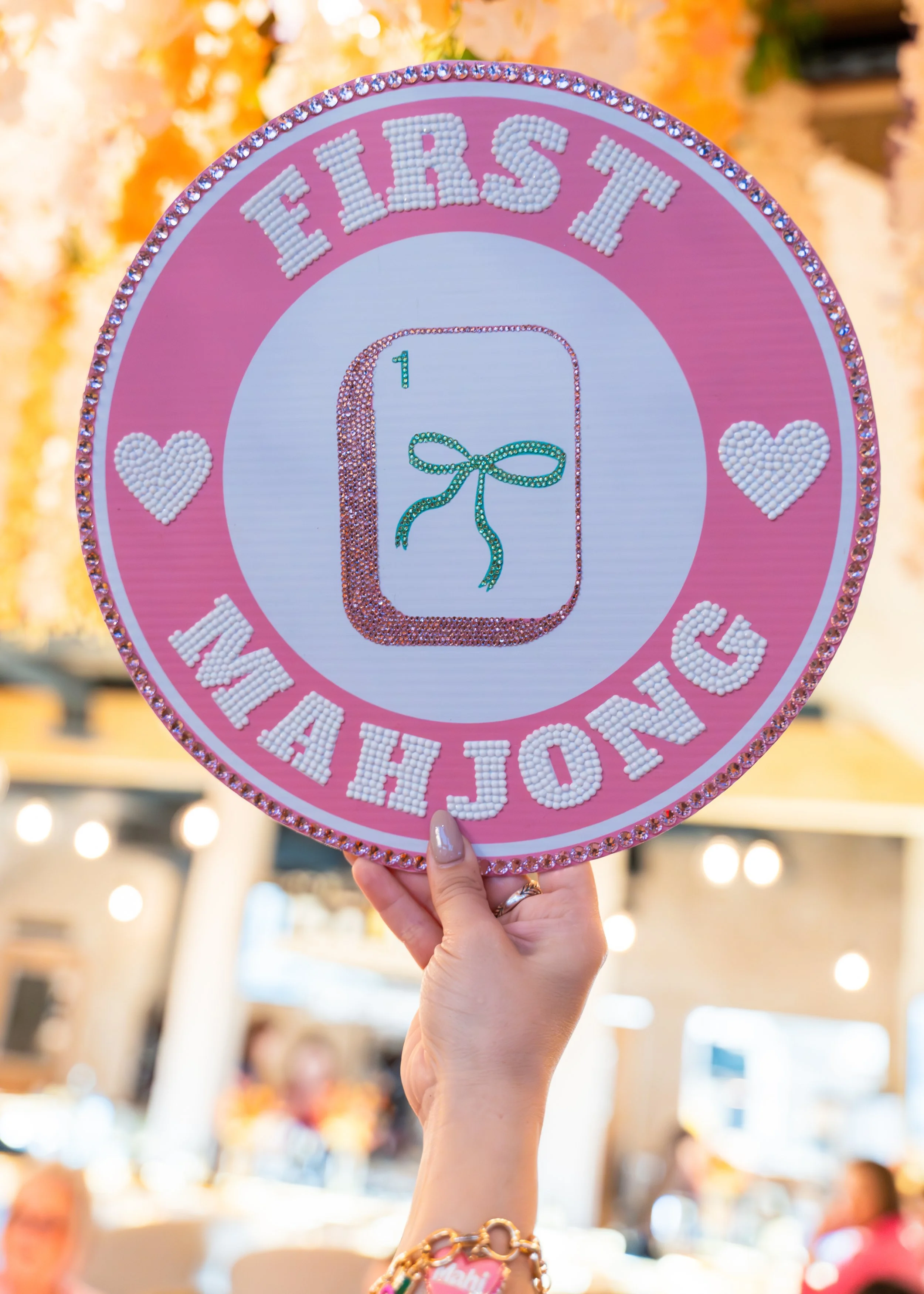 Pink circular sign decorated with rhinestones, displaying the words 'First Year' and a number one with a bow in the center, held by a person with jewelry on their wrist.