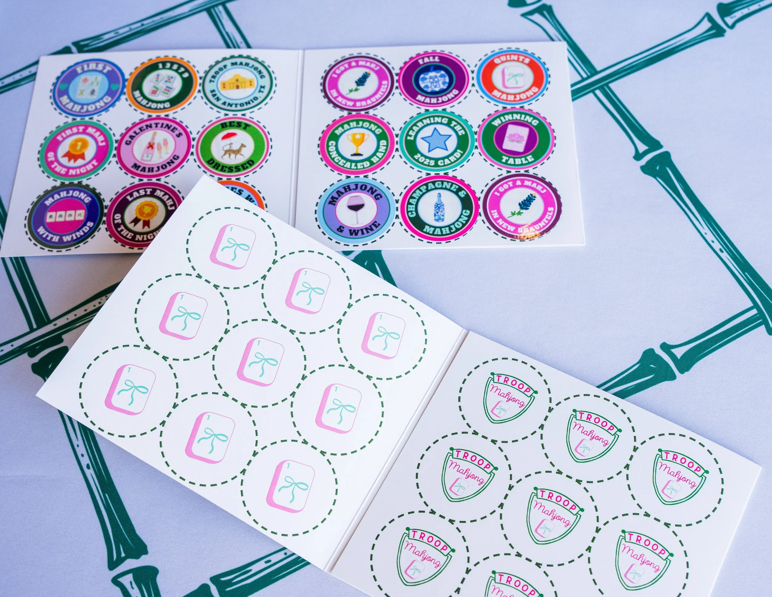 Colorful stickers and labels related to Mahjong game, with some indicating first, last, or best games, and others displaying symbols like a wine glass, a trophy, or a gift box, placed on a leaf-patterned tablecloth.