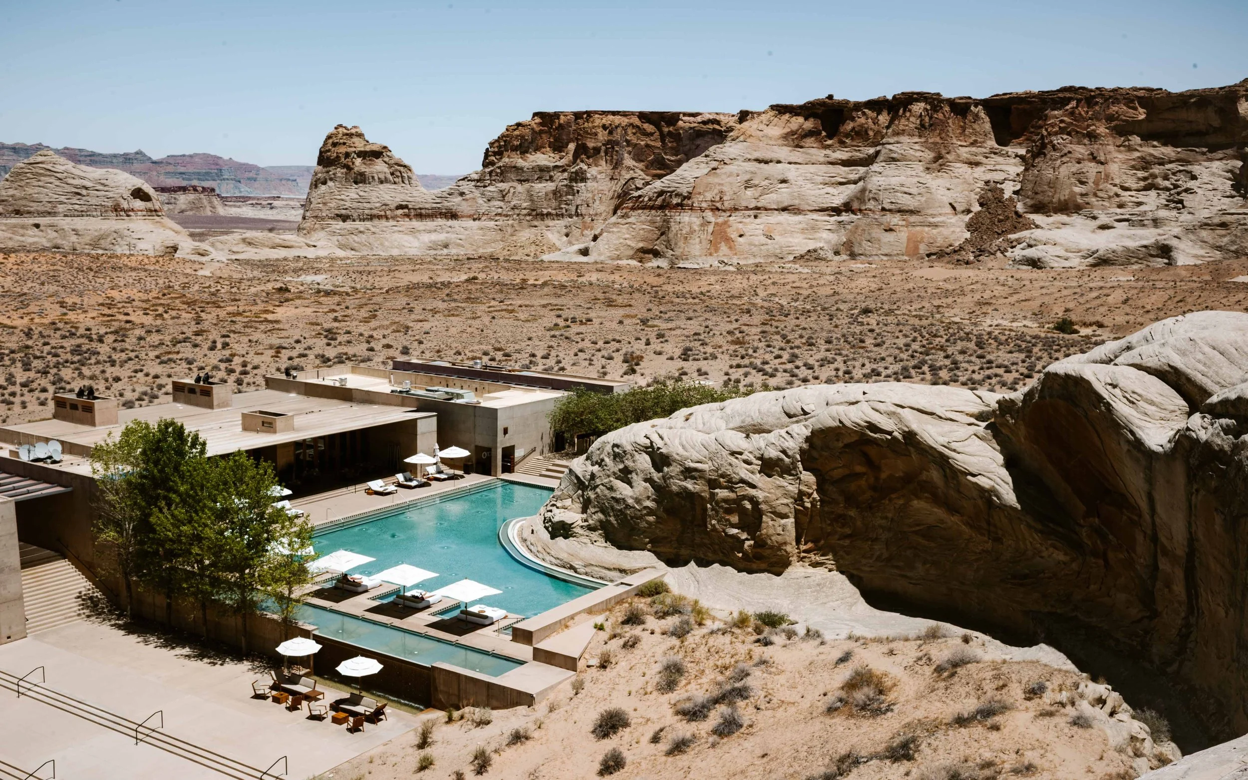 Luxury hotel in a desert landscape with large rock formations in the background, a swimming pool surrounded by lounge chairs and umbrellas, and modern buildings with rooftop terraces.