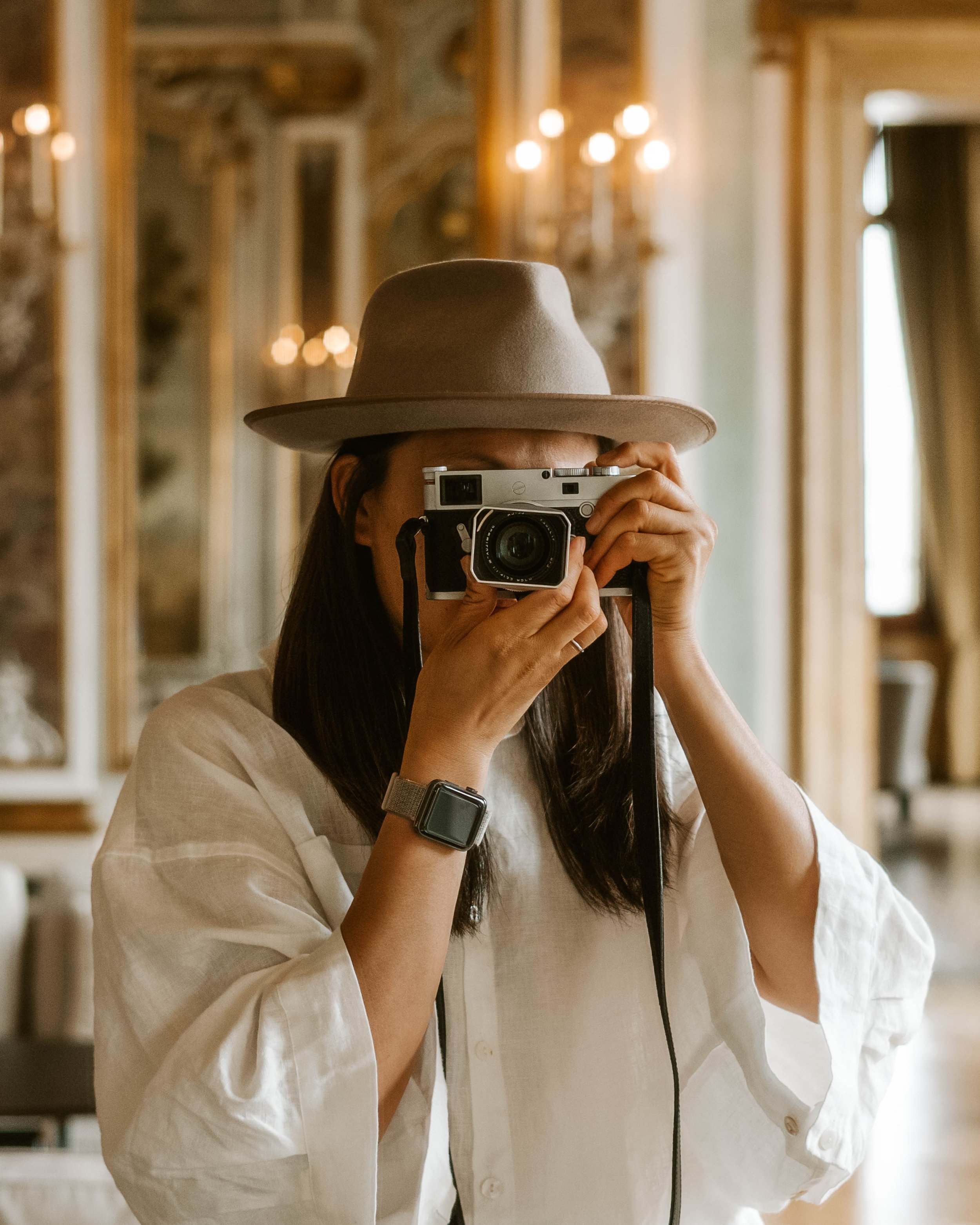 Person taking a photo with a vintage film camera in an elegant room with ornate chandeliers, large mirror, and gold-framed wall decor, wearing a beige fedora and a white shirt.
