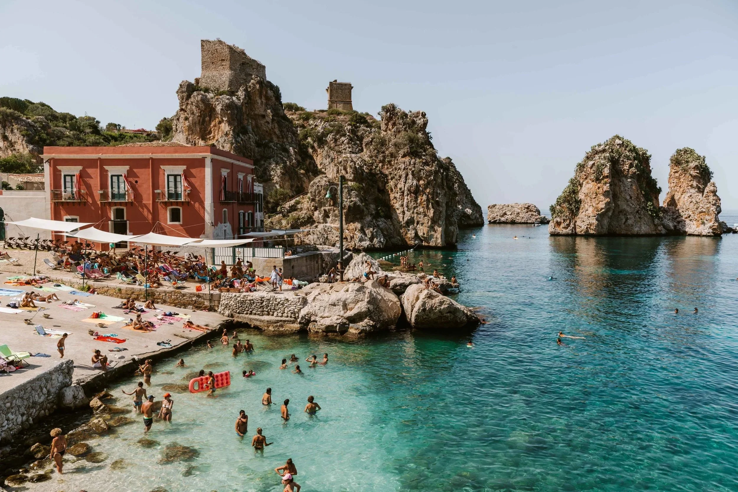 People swimming and sunbathing at a beach with rocky cliffs and historic towers in the background, under a clear sky.