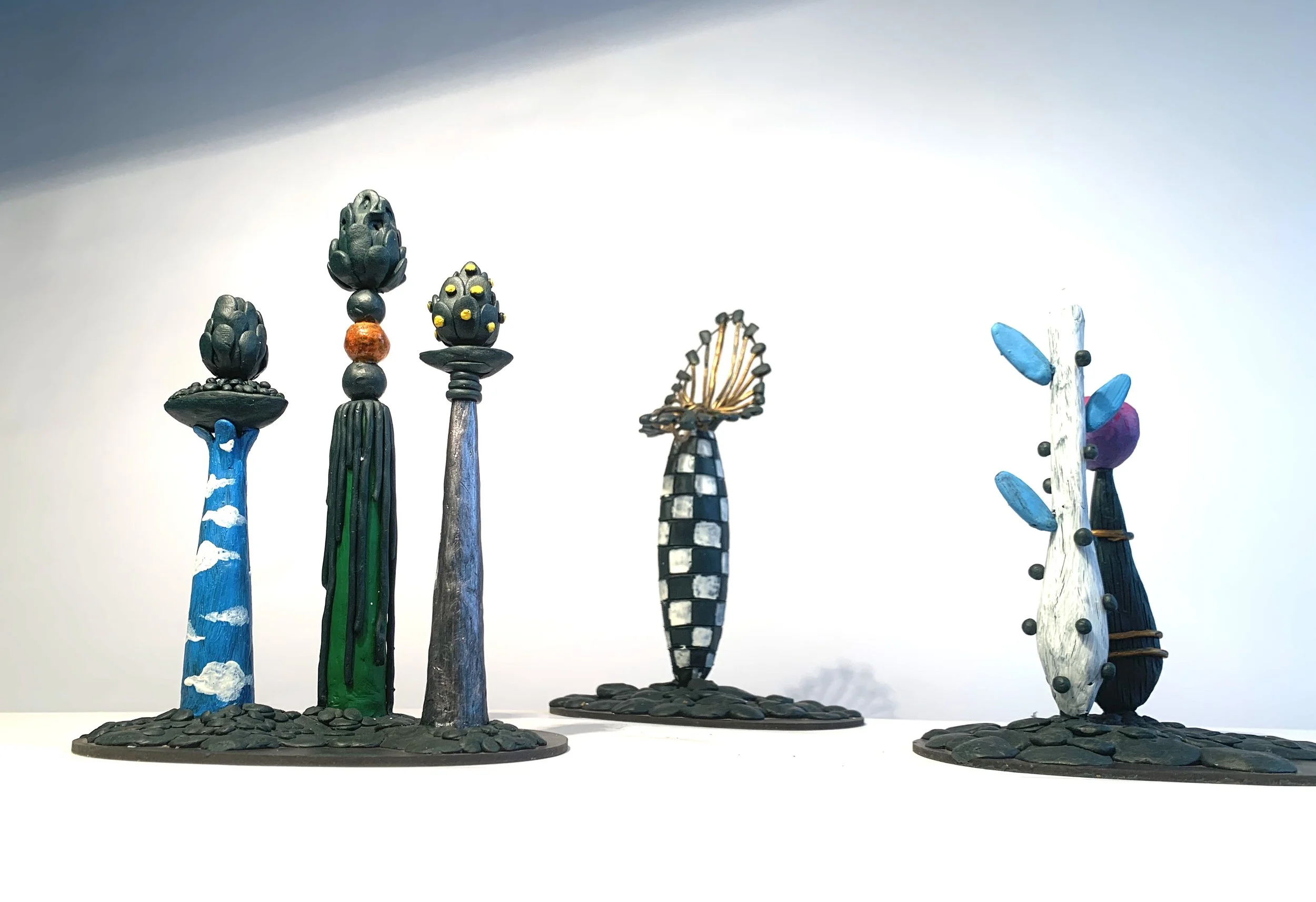 Six colorful, artistic, tree-like sculptures with unique designs and textures displayed on a white surface against a plain background.