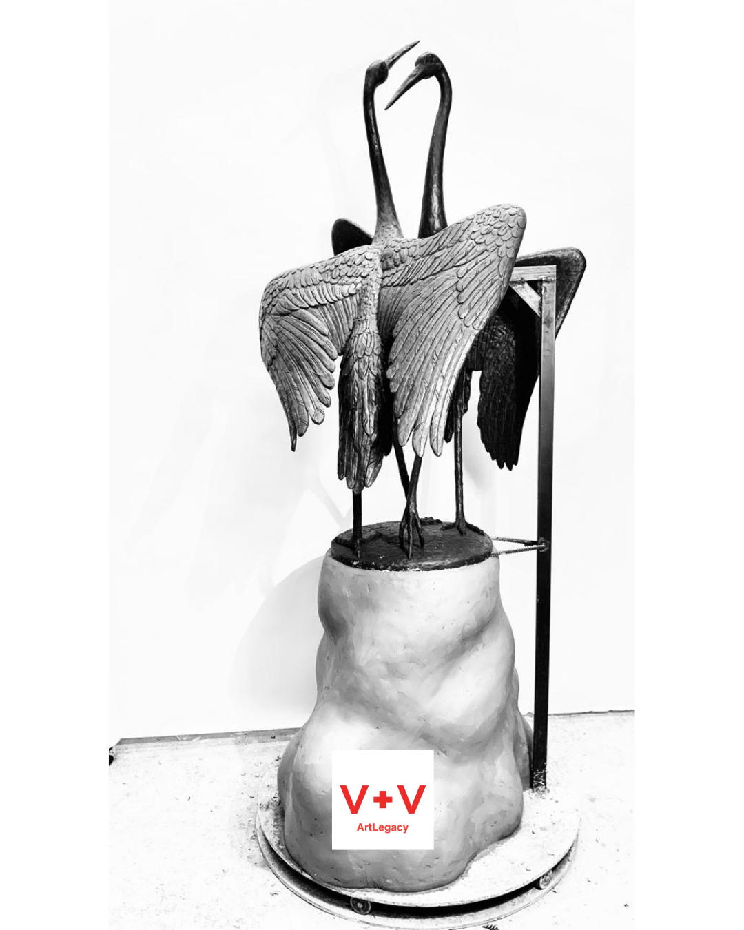 A black and white sculpture of two herons standing on a base, with their wings partially extended, set against a plain white background.