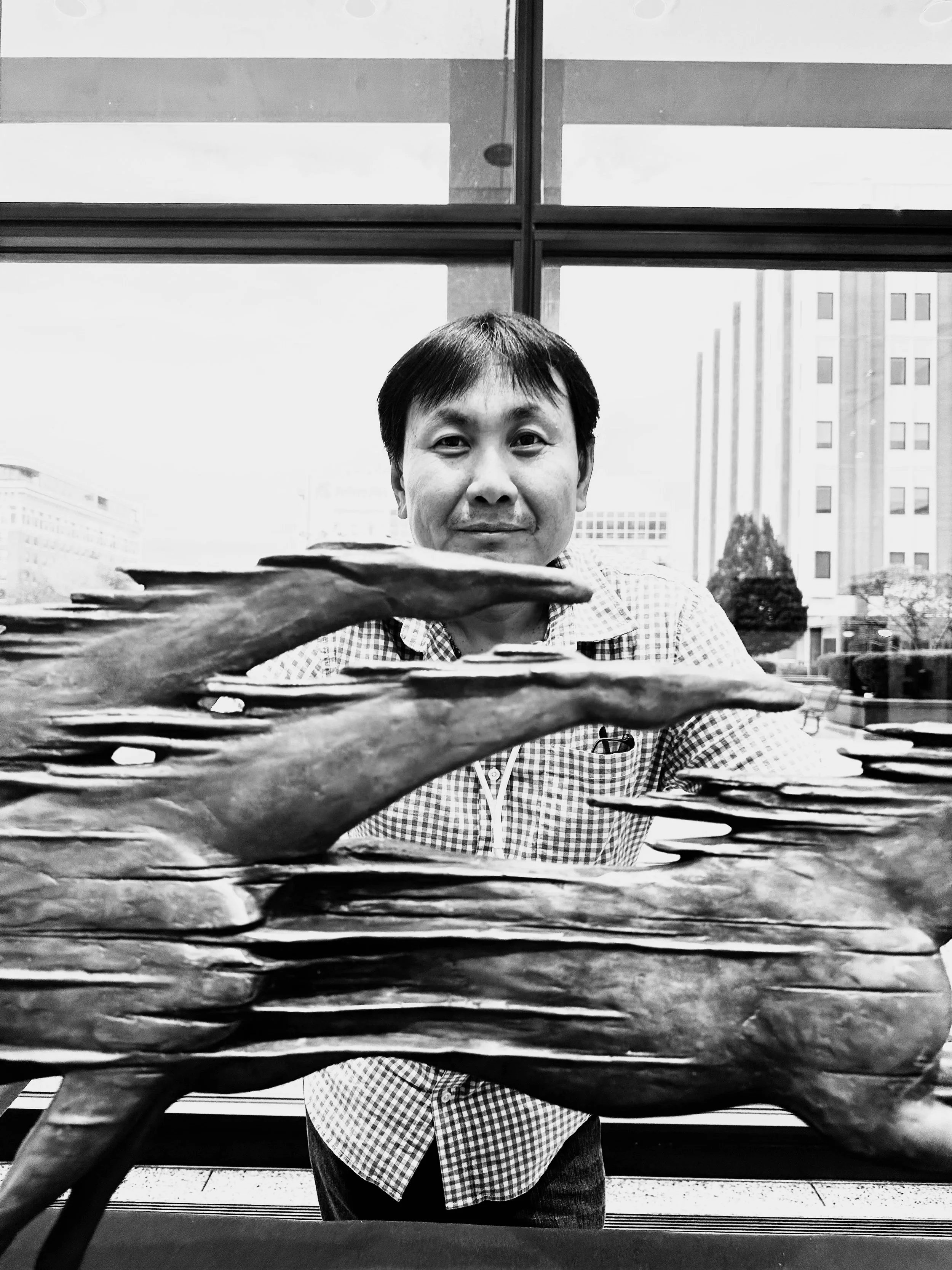 A black and white photo of a man with short hair, possibly of Asian descent, sitting at a table, with a window and city buildings in the background. There are several stacked plates or dishes on the table in the foreground.