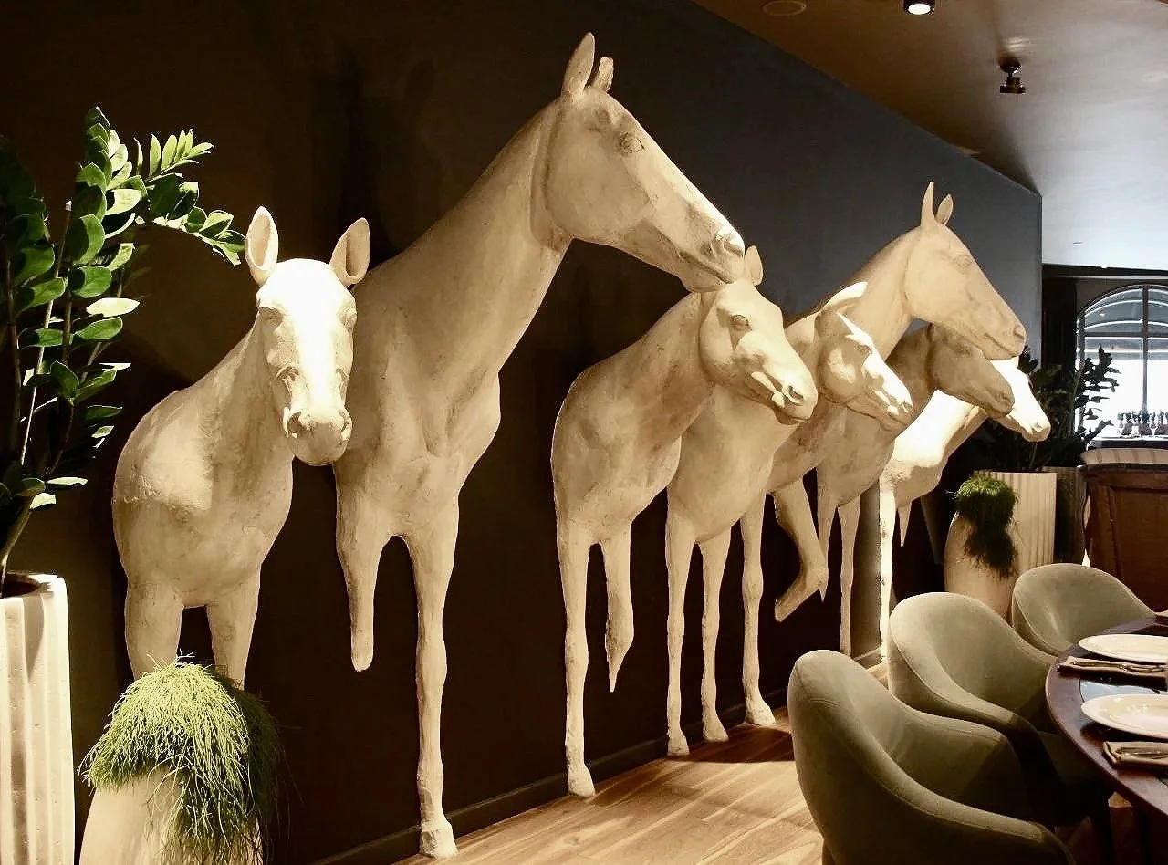 A row of large white horse sculptures mounted on a wall inside a modern restaurant or cafe, with green plants and dining tables in the foreground.