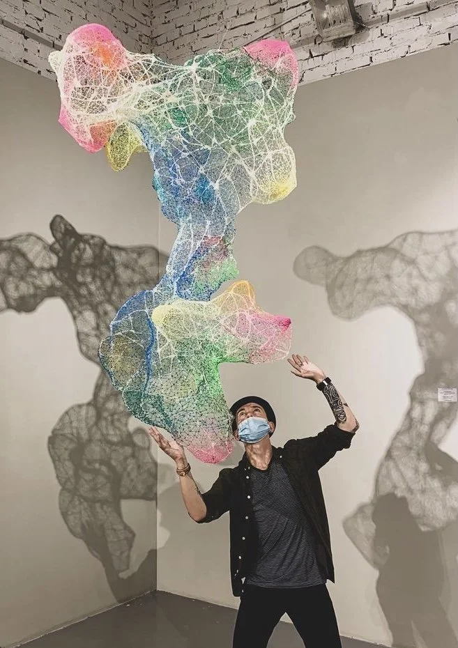 Person wearing a mask stands beneath a large, colorful, abstract wire sculpture creating shadow shadows on the wall.