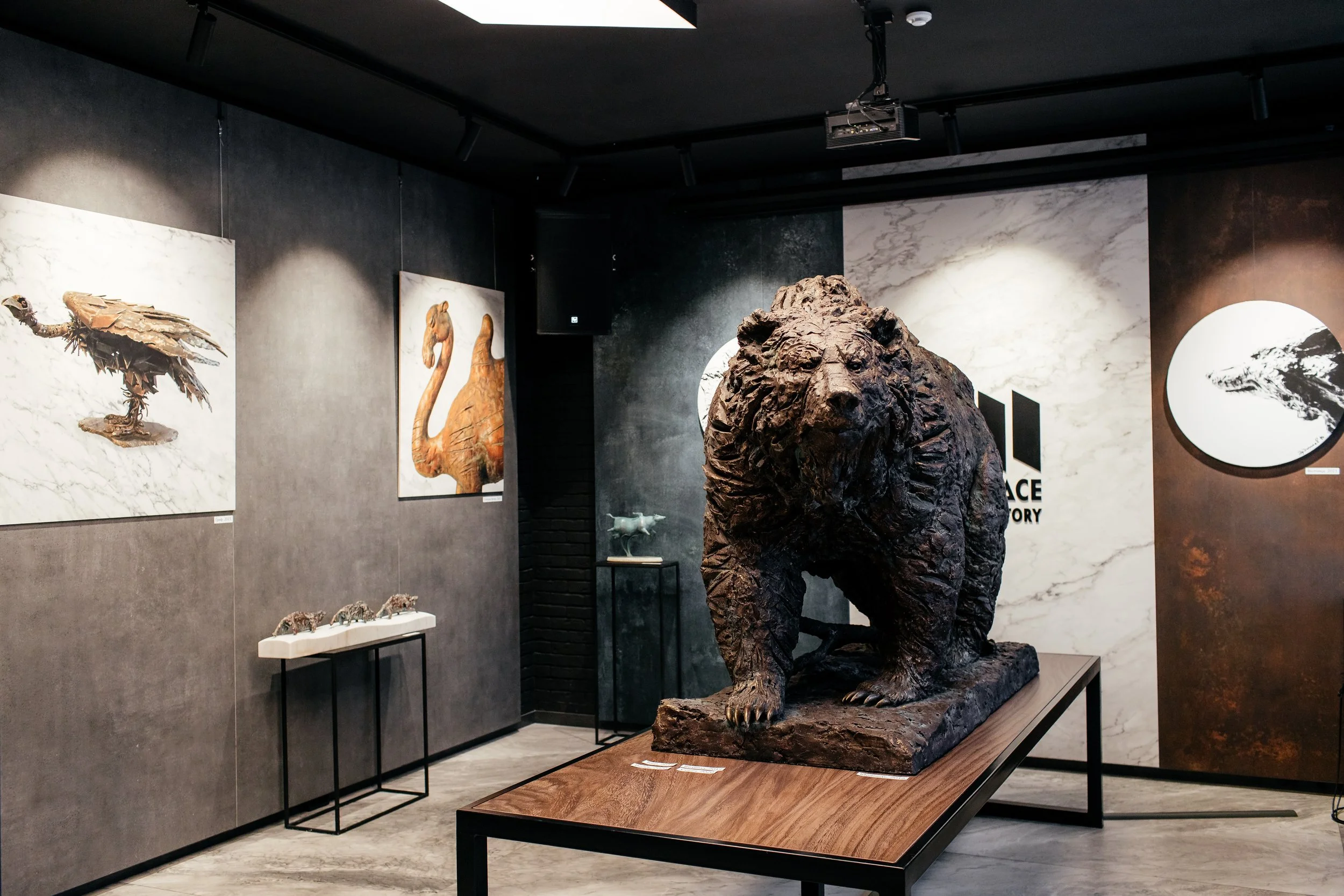 A bronze sculpture of a lion on display in an art gallery with black walls, featuring paintings of various animals on the wall behind it.