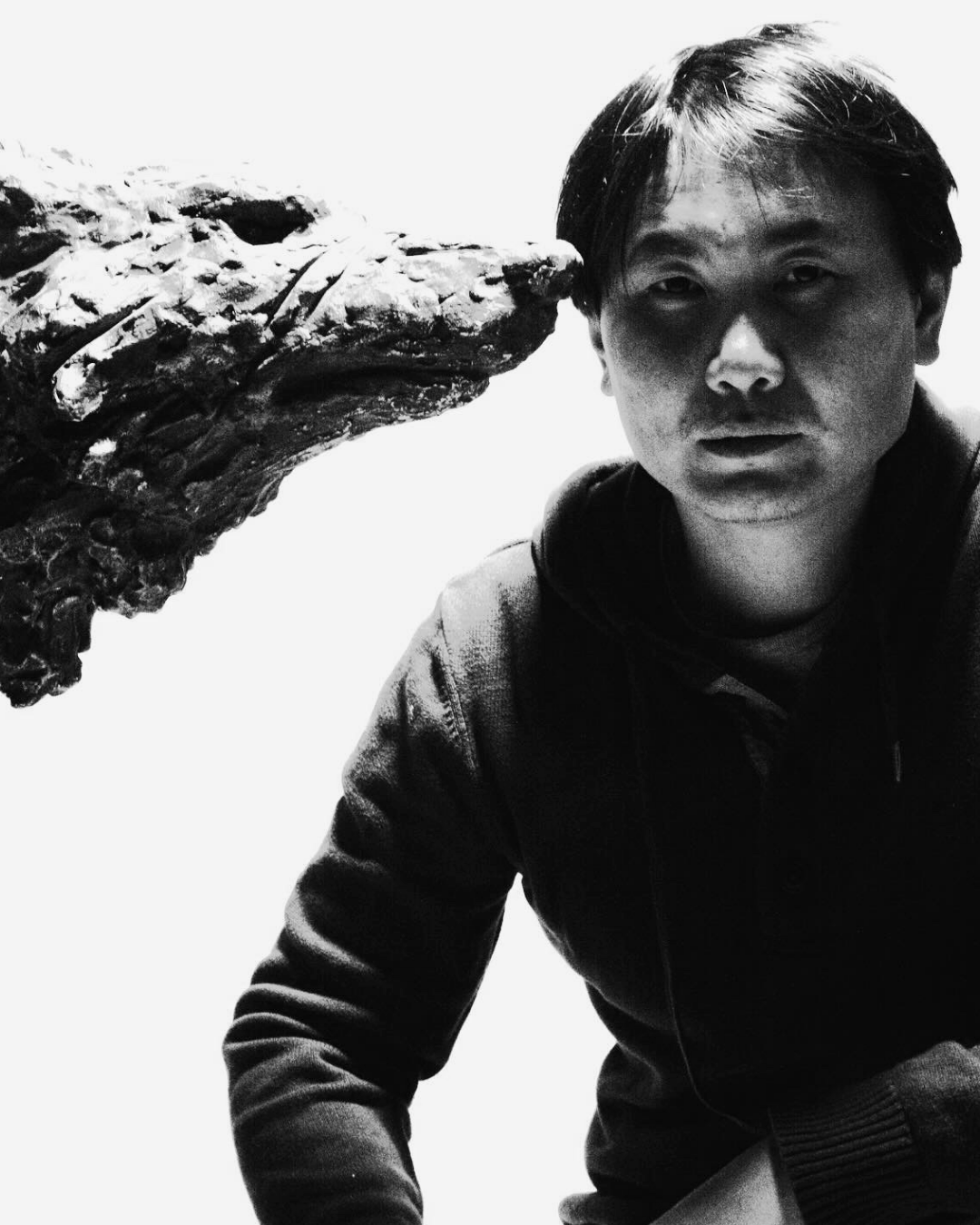 A black and white photo of a young man with medium-length hair looking directly at the camera, with a large crocodile head positioned close to his face on the left side of the image.