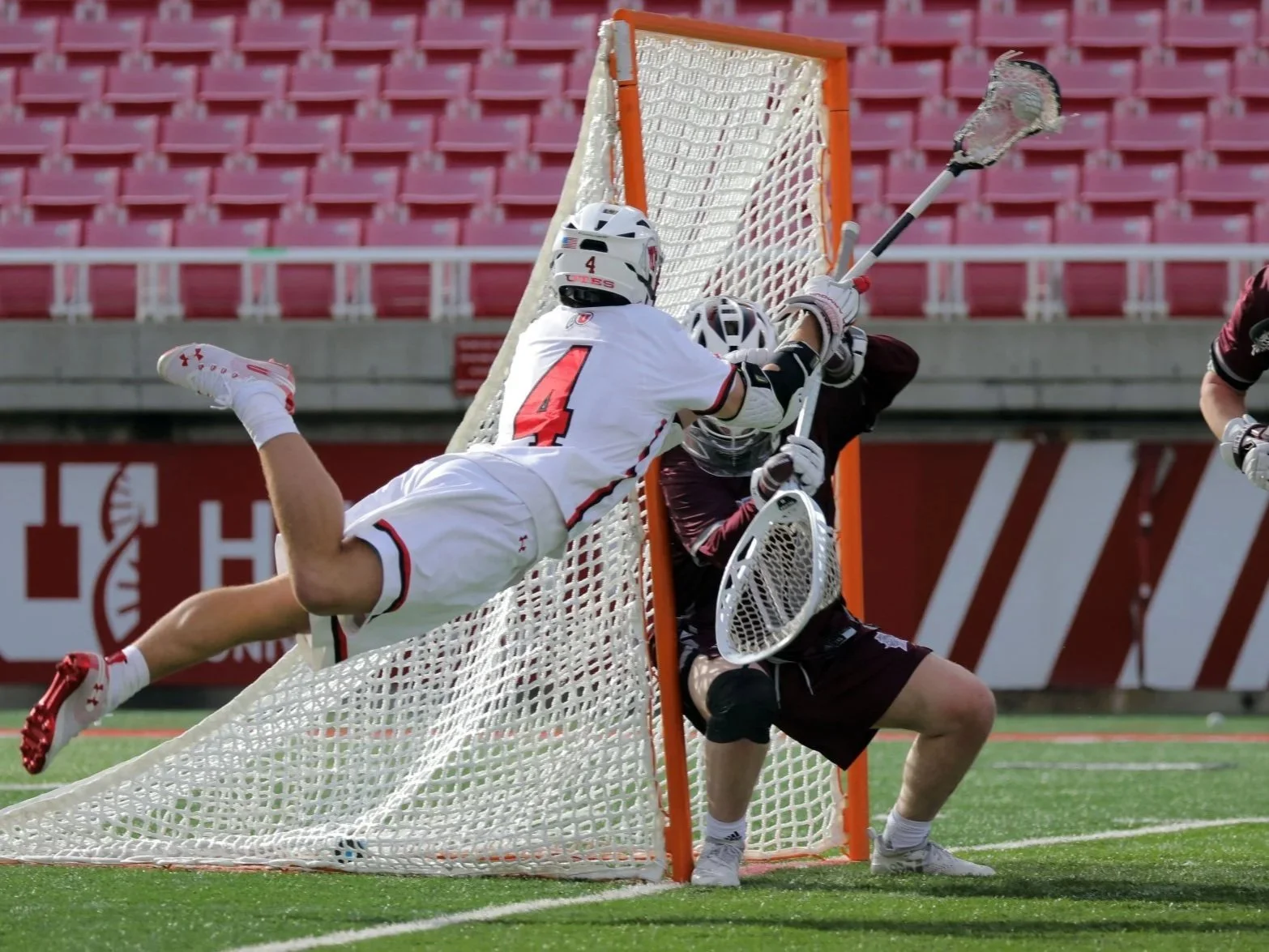 Lacrosse players defend a goal, with one player in a white uniform attempting to score and a goalie in a dark uniform preparing to block the shot, on a green field with red stadium seats in the background.