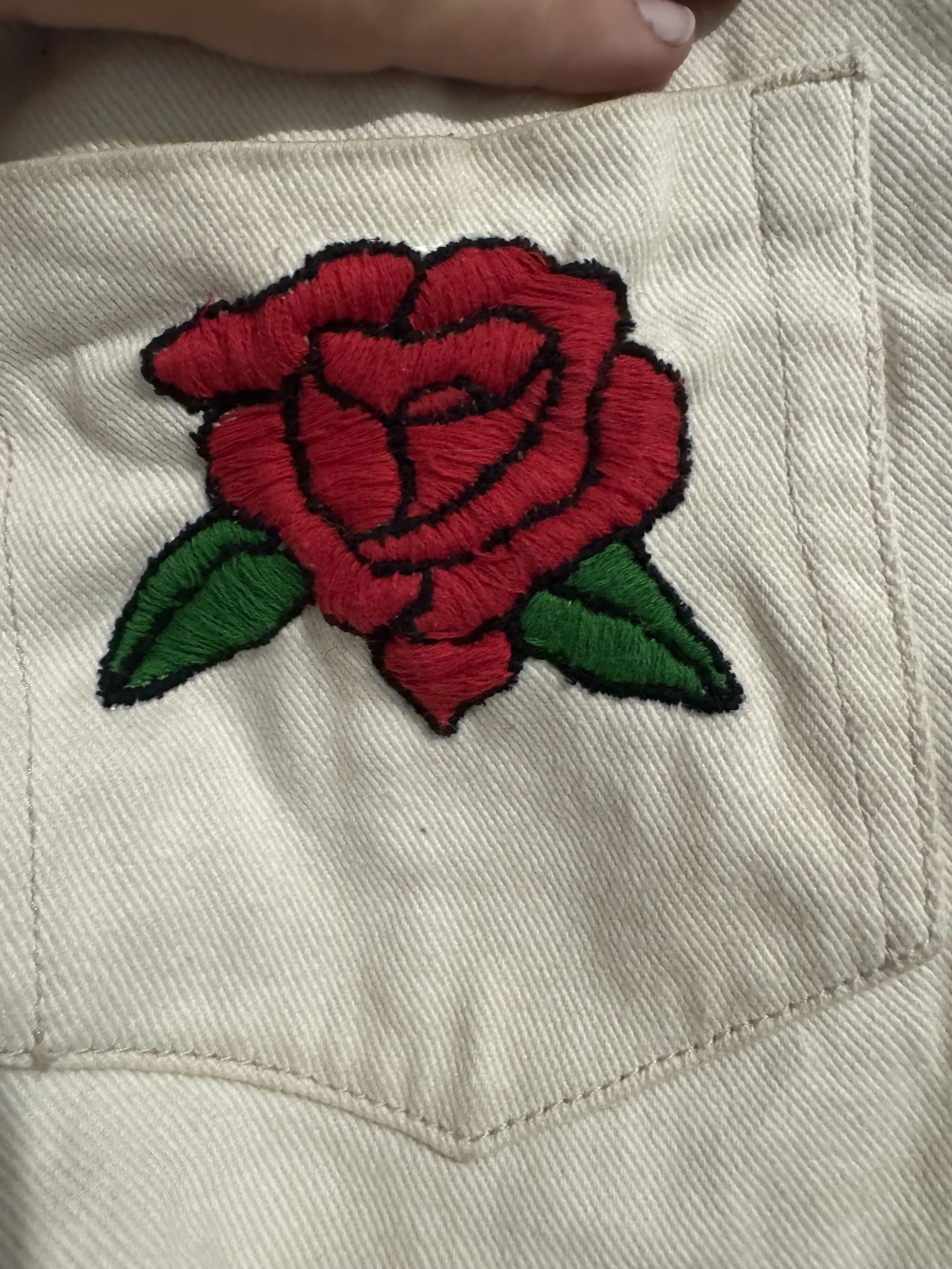 Close-up of a white denim shorts with a stitched embroidered red rose and two green leaves on the side of a pocket.
