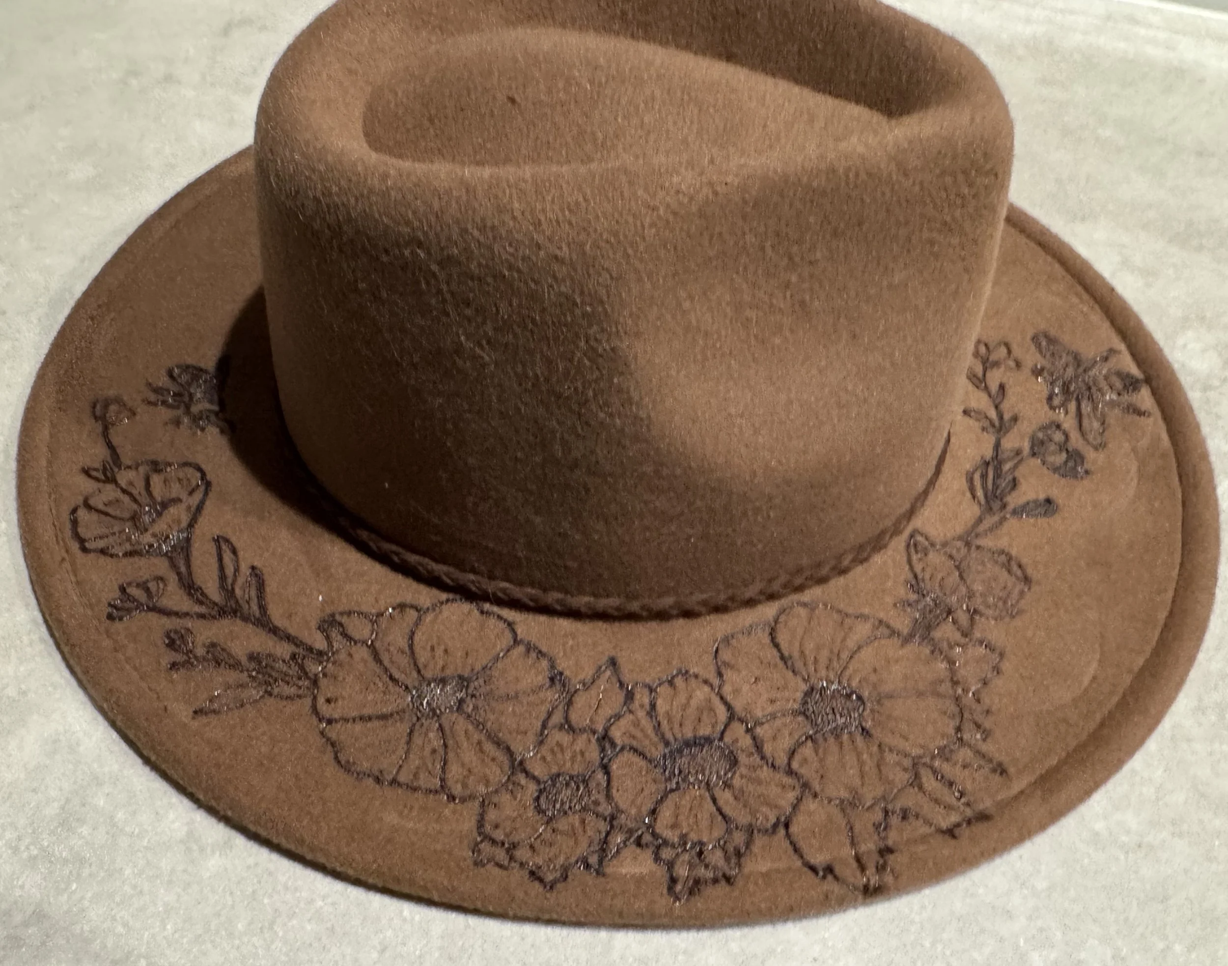 Close-up of a brown suede fedora hat with an floral designs burnt into it.