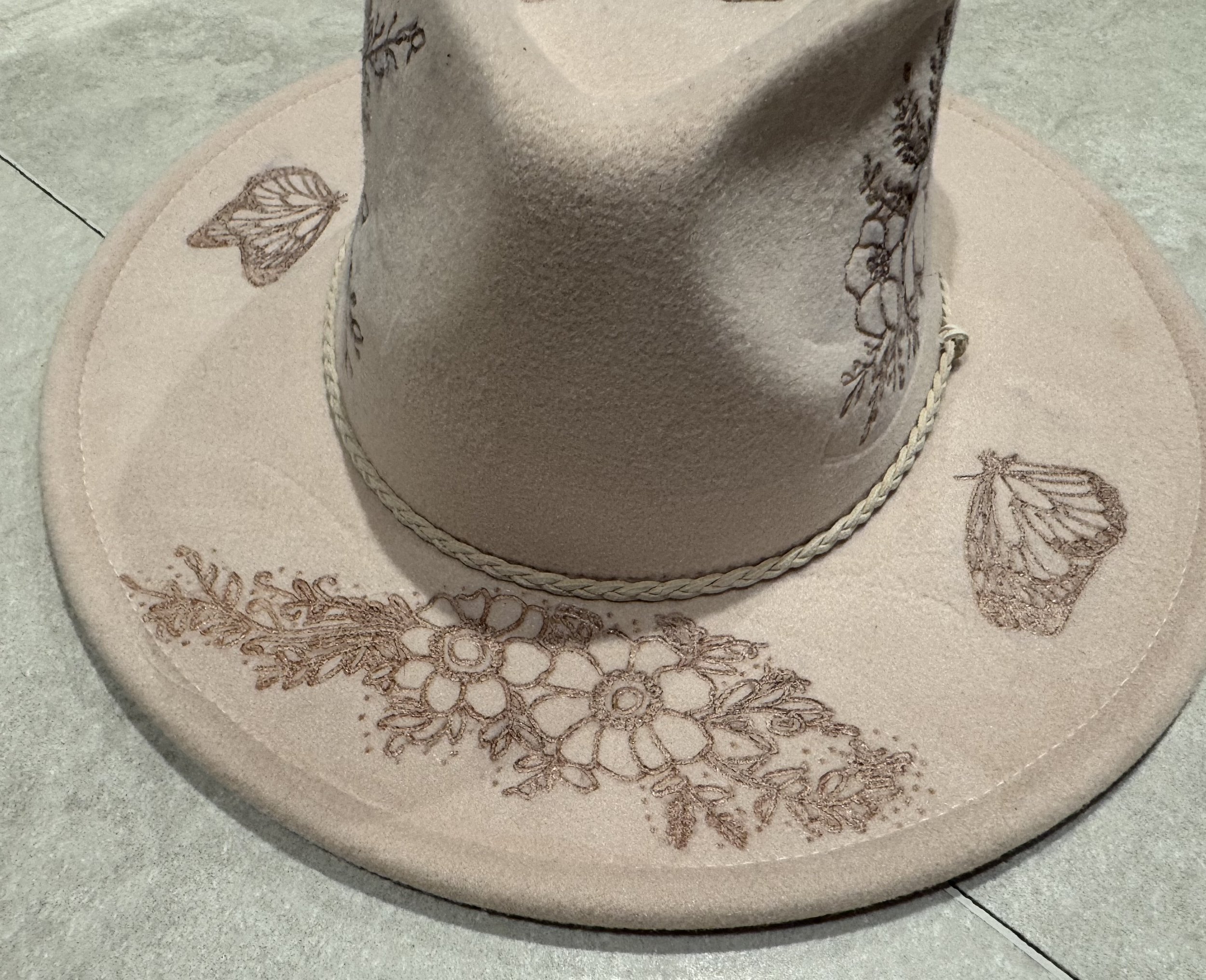 Close-up of a beige cowboy hat with floral designs and butterflies burnt into it.