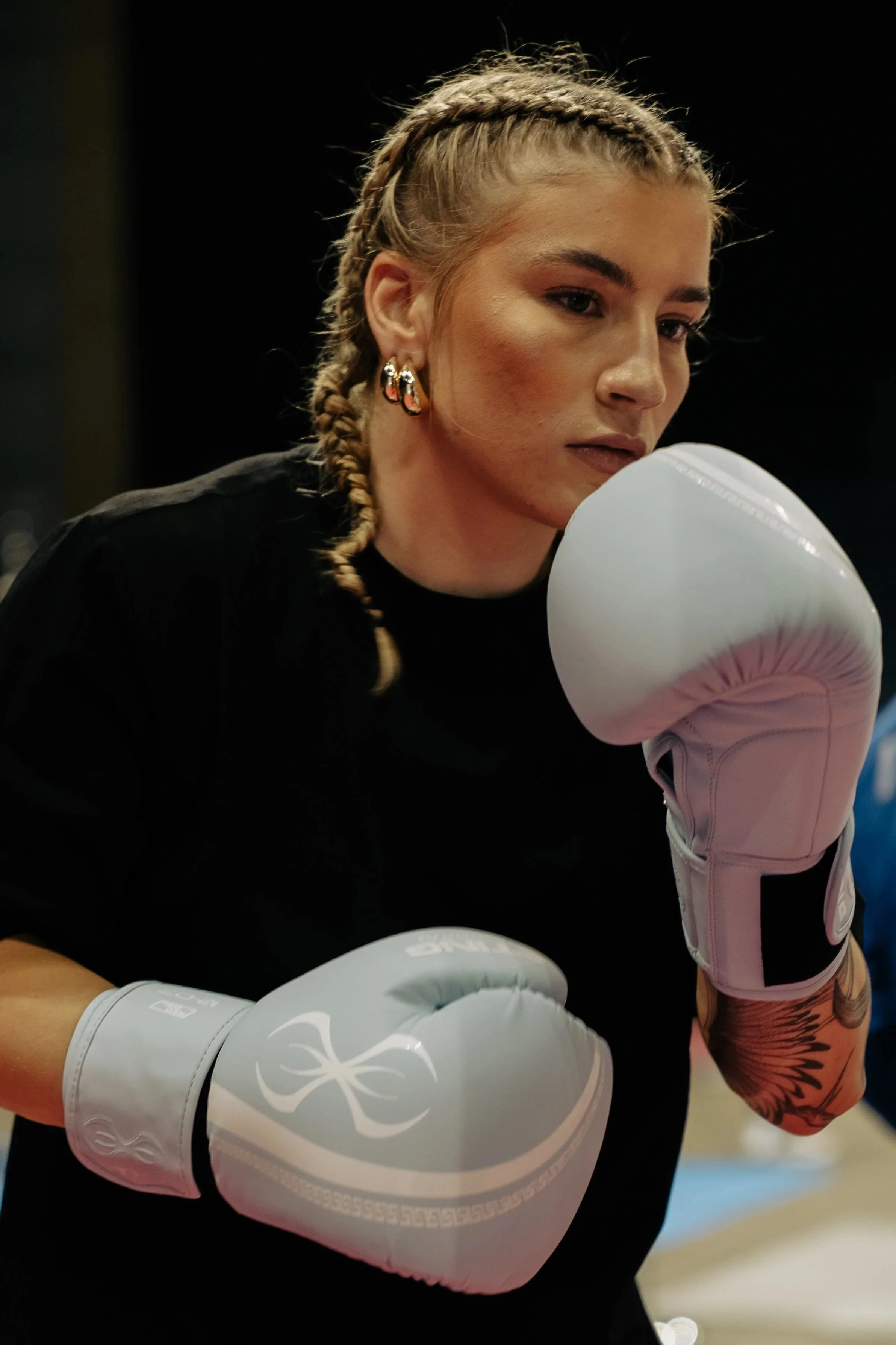 A young woman with braided blonde hair and hoop earrings wearing boxing gloves is in a boxing gym.