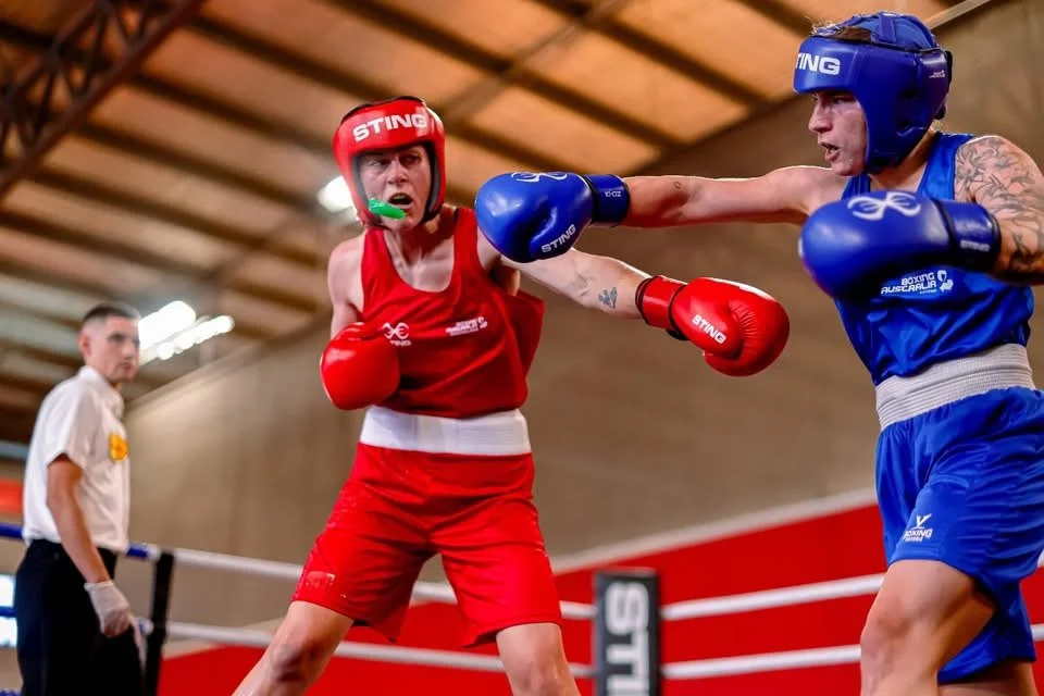 A female boxer in red gear being punched by a male boxer in blue gear inside a boxing ring.