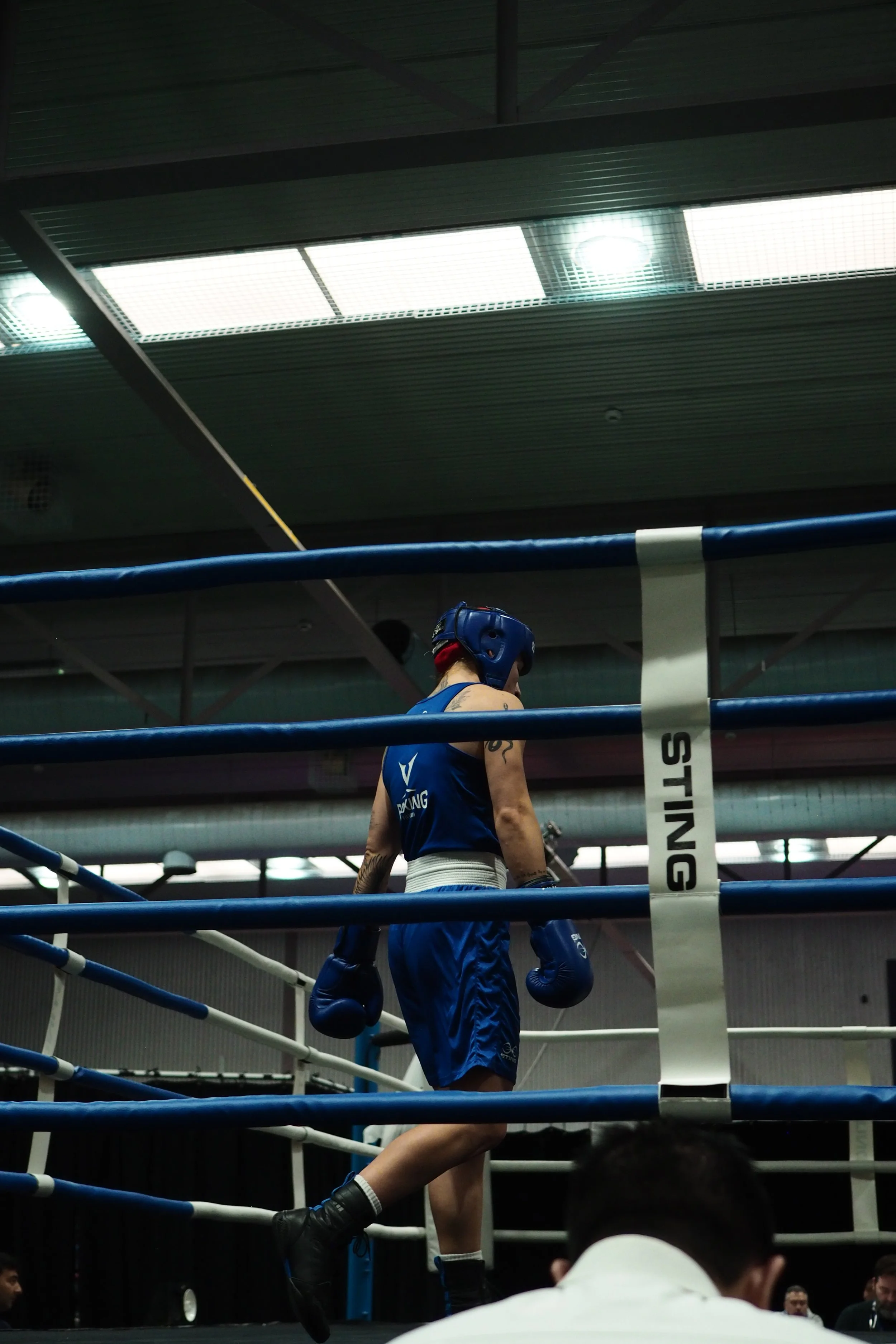 A boxer in blue gear and gloves standing inside a boxing ring, with a person in white in the foreground and a group of people in the background, under bright overhead lights.