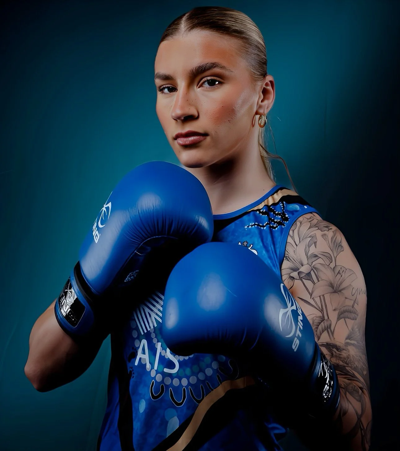 A female boxer in blue gloves and a blue athletic top, with a tattoo sleeve on her left arm, posing with fists raised against a dark background.
