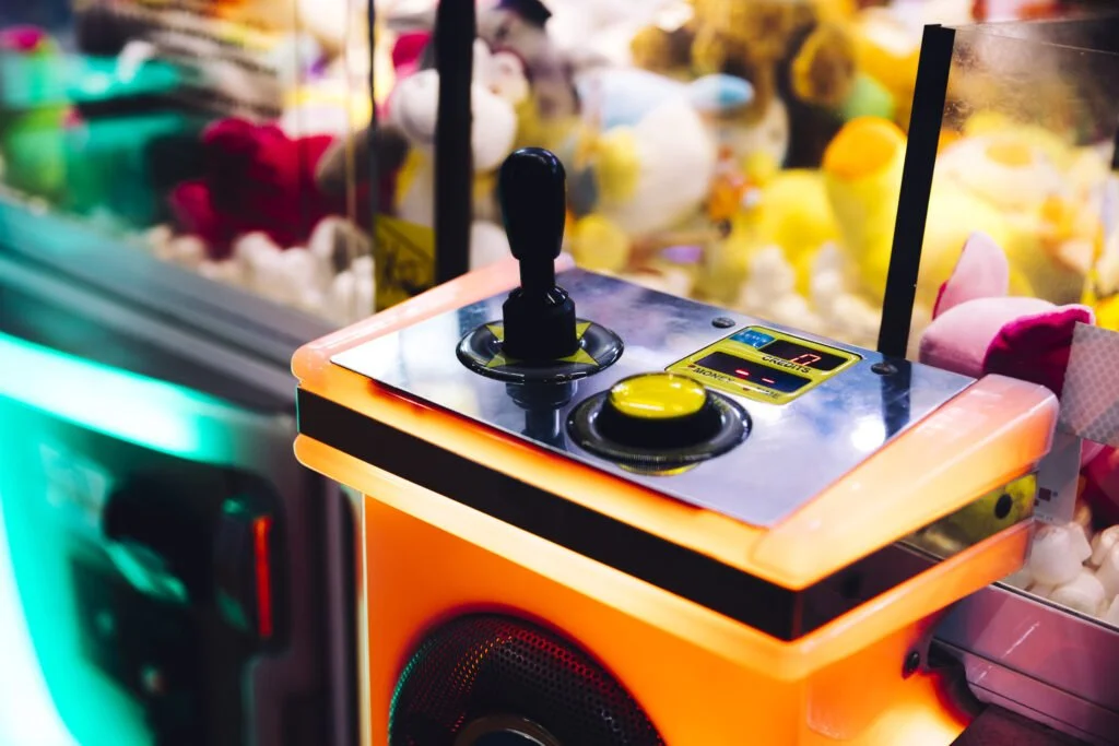 Close-up of a claw machine control panel with a joystick and a yellow button, surrounded by plush toys inside the claw arcade game.