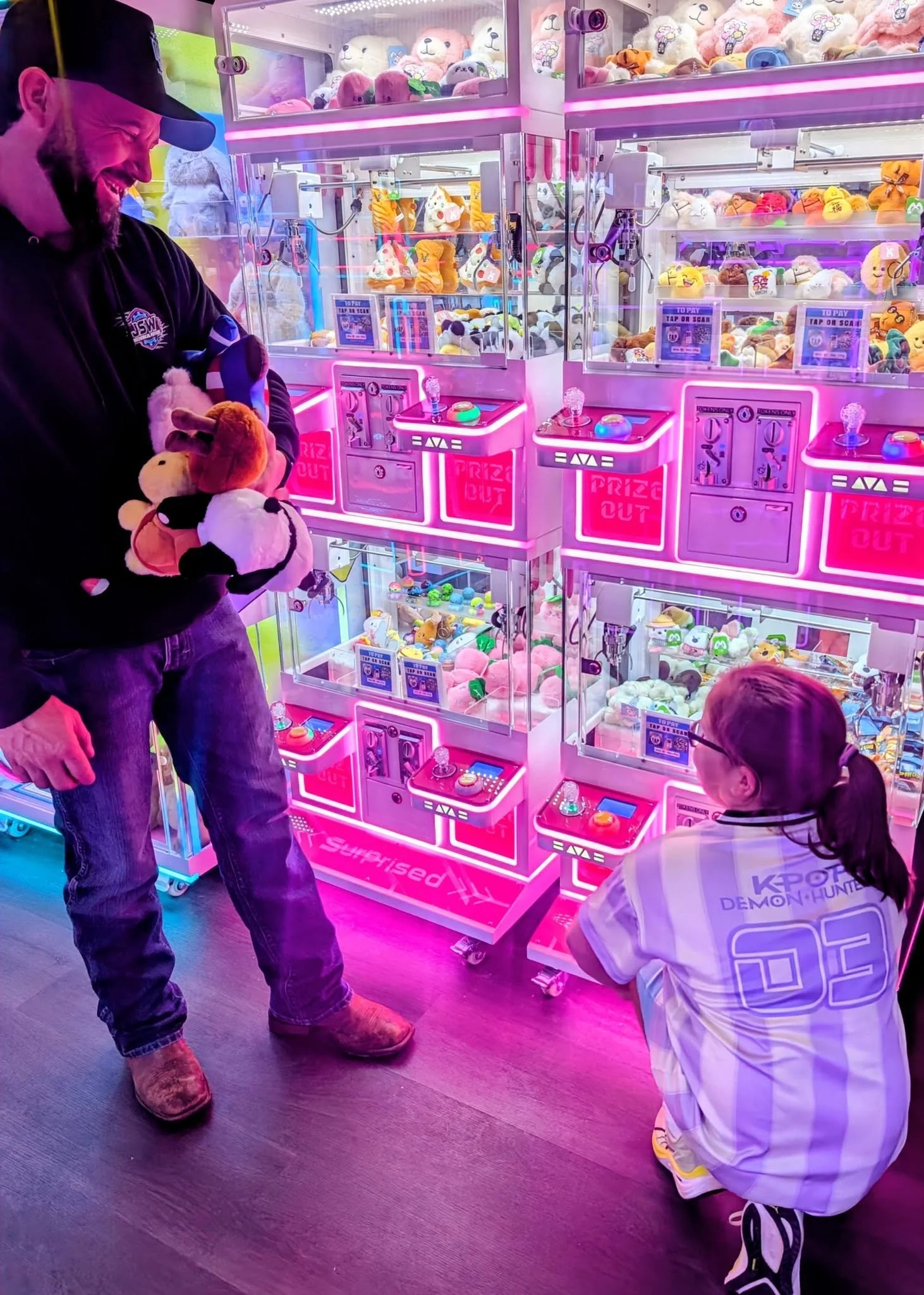 A man wearing a black jacket and cap, holding plush toys, and a girl in a striped shirt and glasses, looking at a colorful claw machine filled with stuffed toys at an arcade.