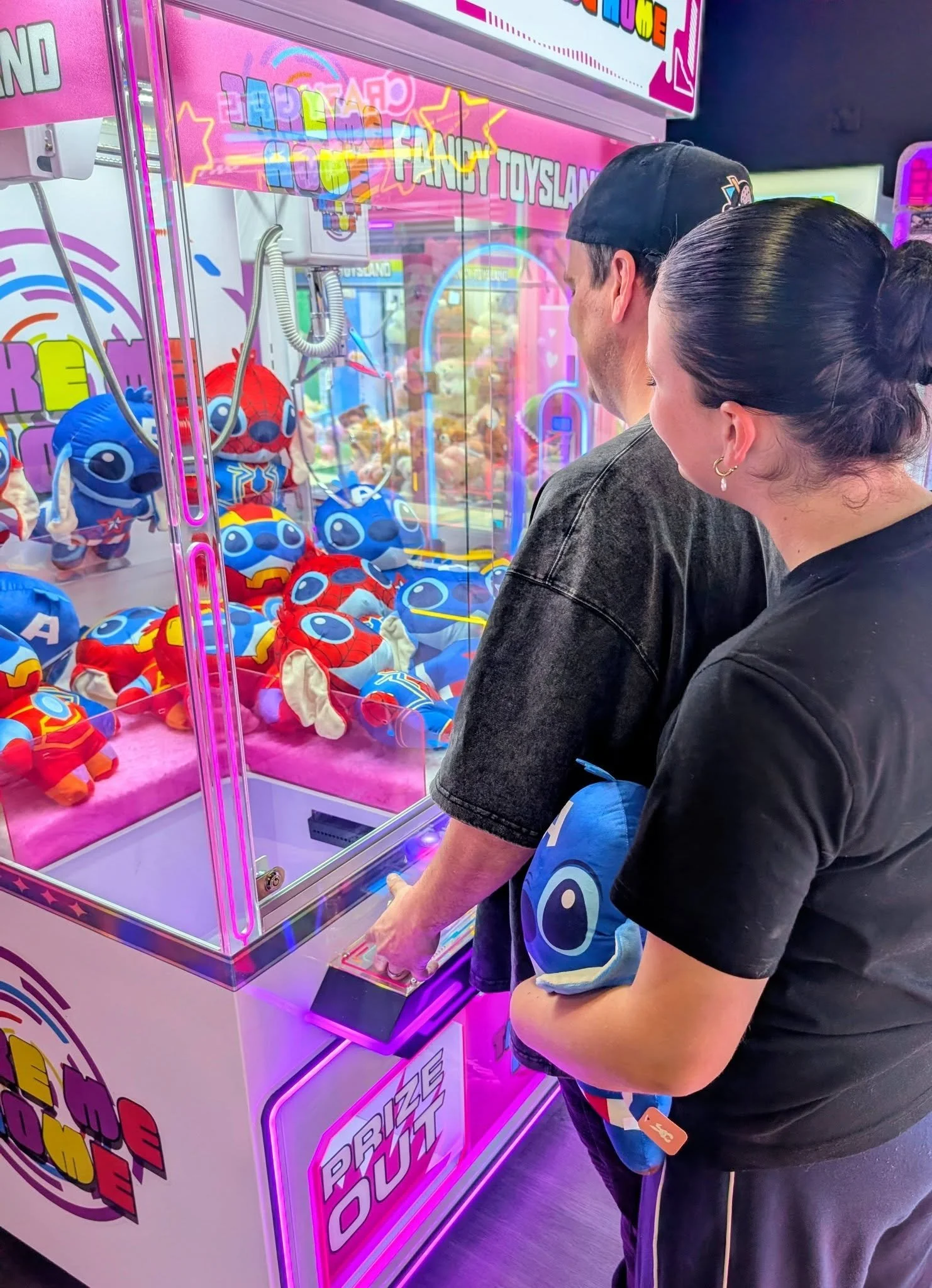 Two people playing a claw machine game at an arcade, filled with colorful plush toys, with neon pink and purple lights and fun signage.