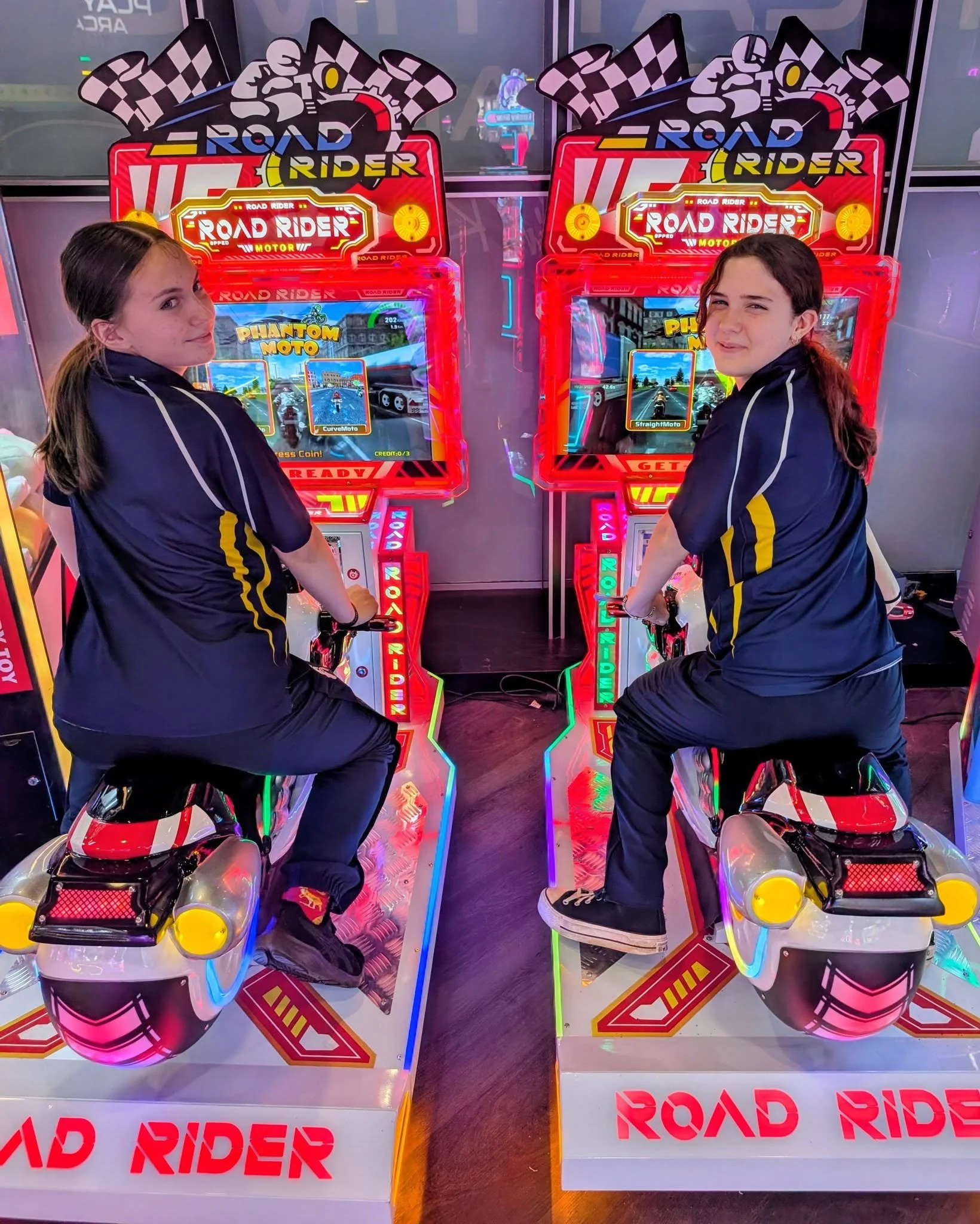Two girls sitting on arcade motorcycle riding simulators in front of a colorful neon arcade machine titled 'Road Rider'.