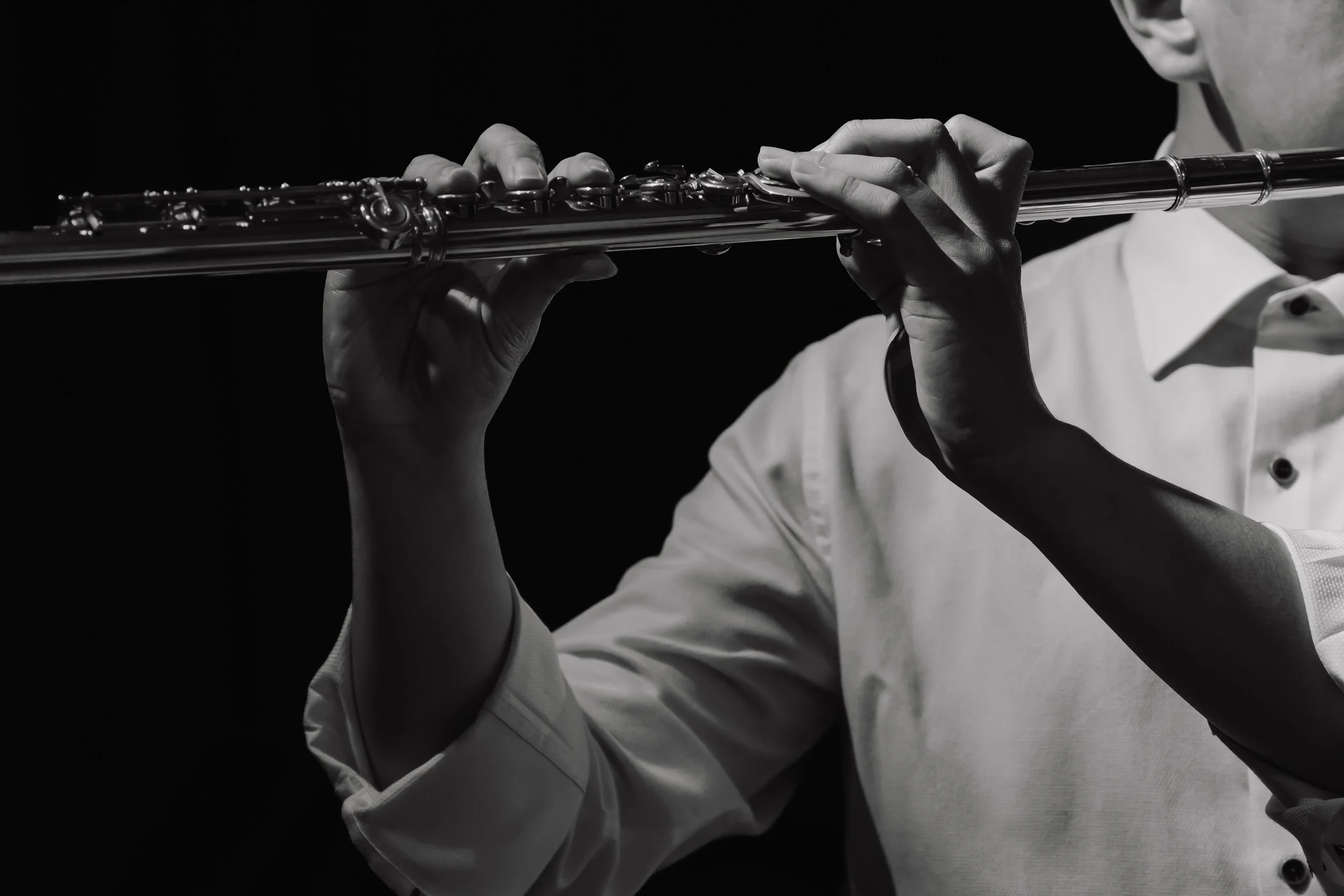 A person playing the flute, wearing a white shirt, captured in black and white against a dark background.