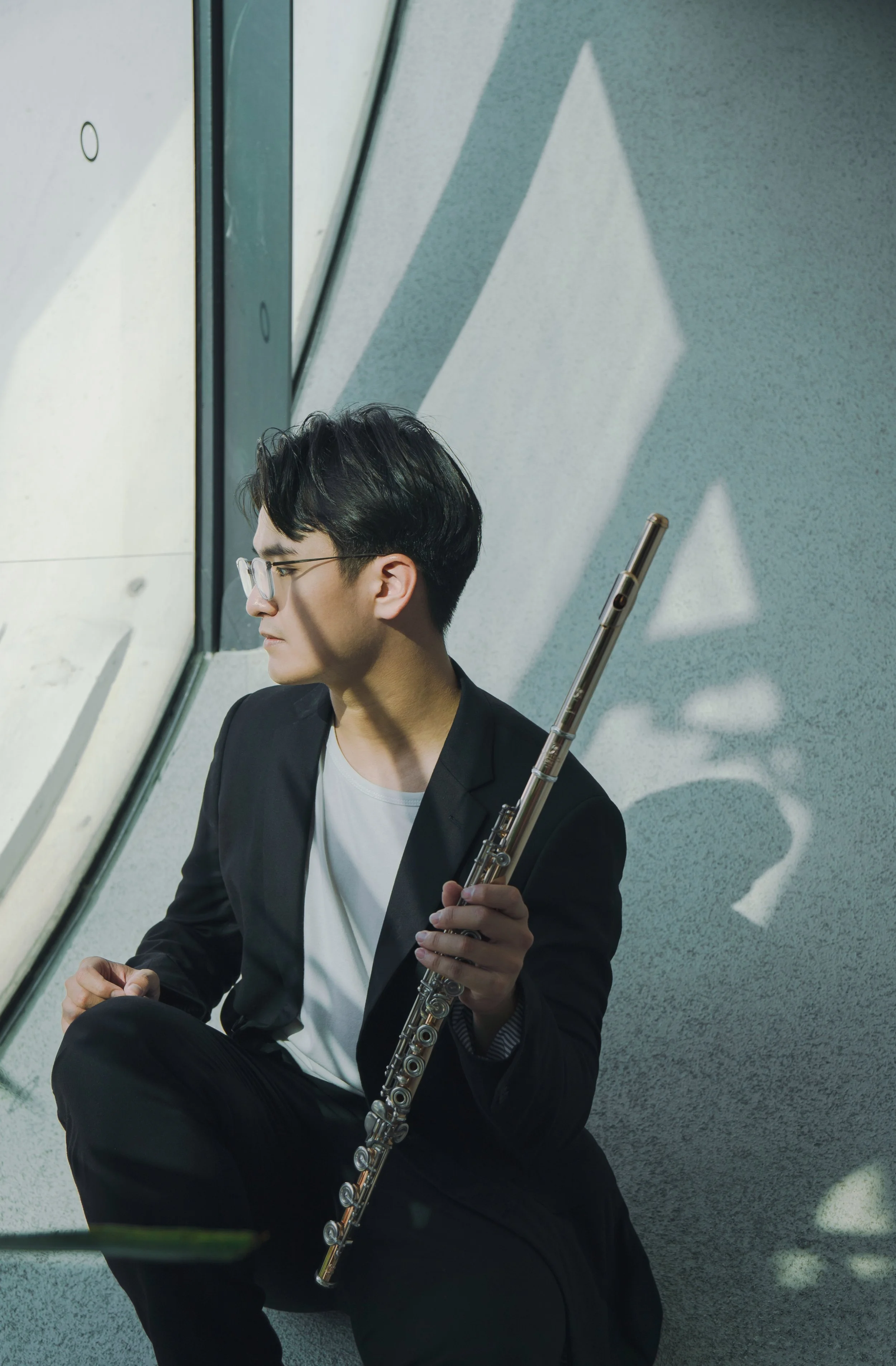 A young man in glasses and a black blazer sits on the floor in front of a large window, holding a flute and looking downward. His shadow creates a geometric pattern on the wall behind him.