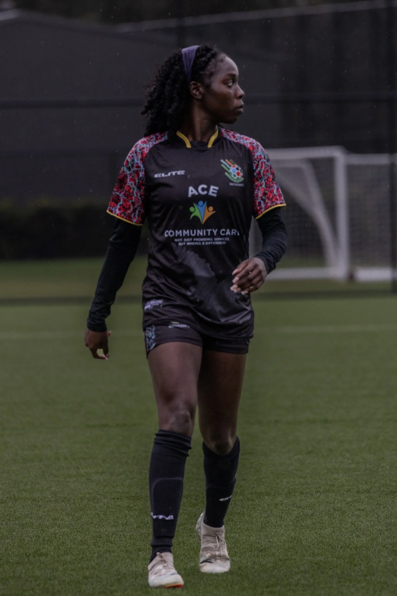Female soccer player in black and red uniform walking on a grass field, with a soccer goal in the background.