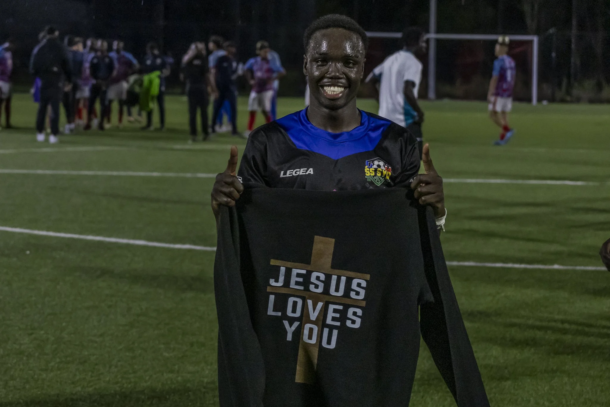 A smiling young man in a black and blue sports jersey holding a sign with the words 'JESUS LOVES YOU' written on it, standing on a soccer field during evening or night, with a group of people in the background.