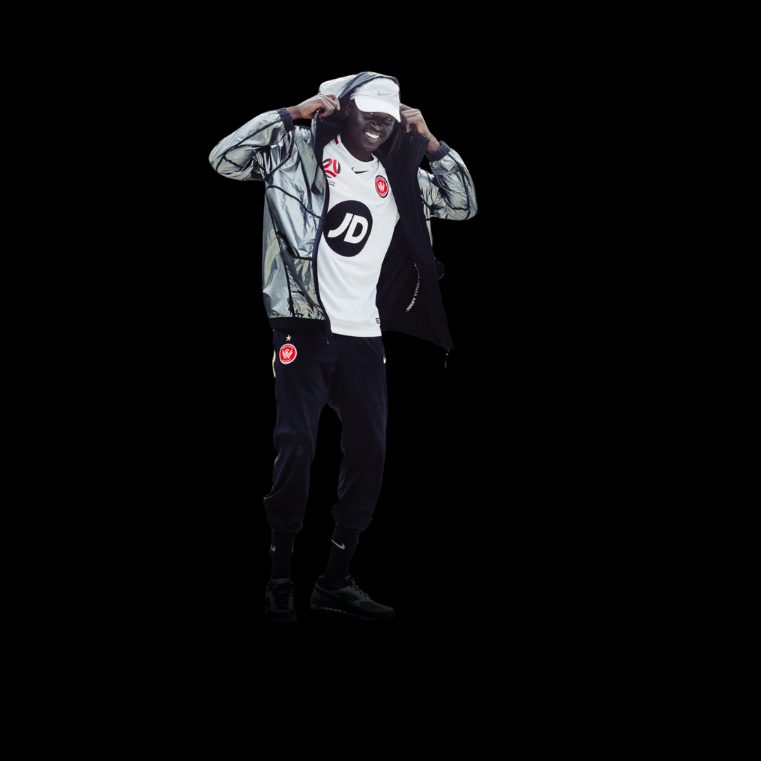 A man standing against a black background, wearing a white sports shirt, black pants, black sneakers, a gray and black jacket, and a white cap, smiling and adjusting his jacket.