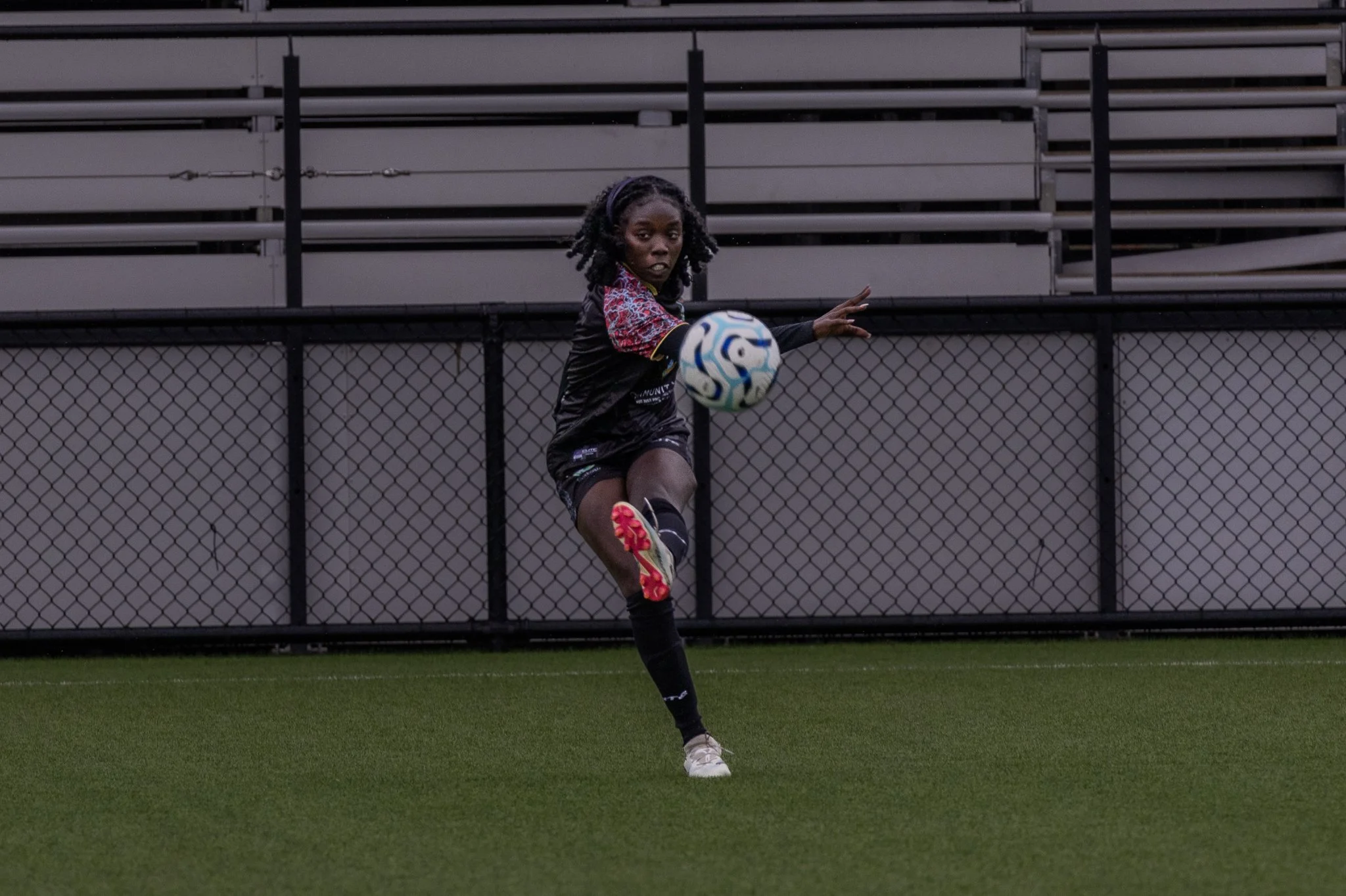 A young woman playing soccer on a field during daytime. She is in mid-action, kicking a soccer ball, with her right leg extended and her arms out to the sides. She has curly hair and is wearing sports clothing.