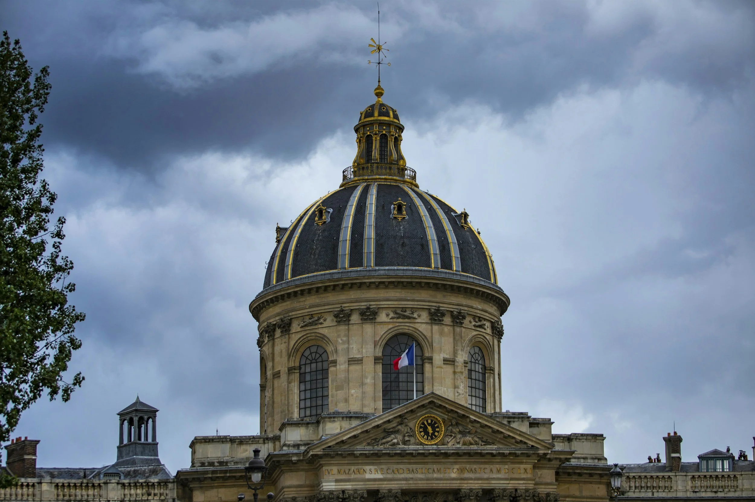 Coupole ornée de dorures sur un bâtiment historique, avec un drapeau français en bas, ciel nuageux en arrière-plan.