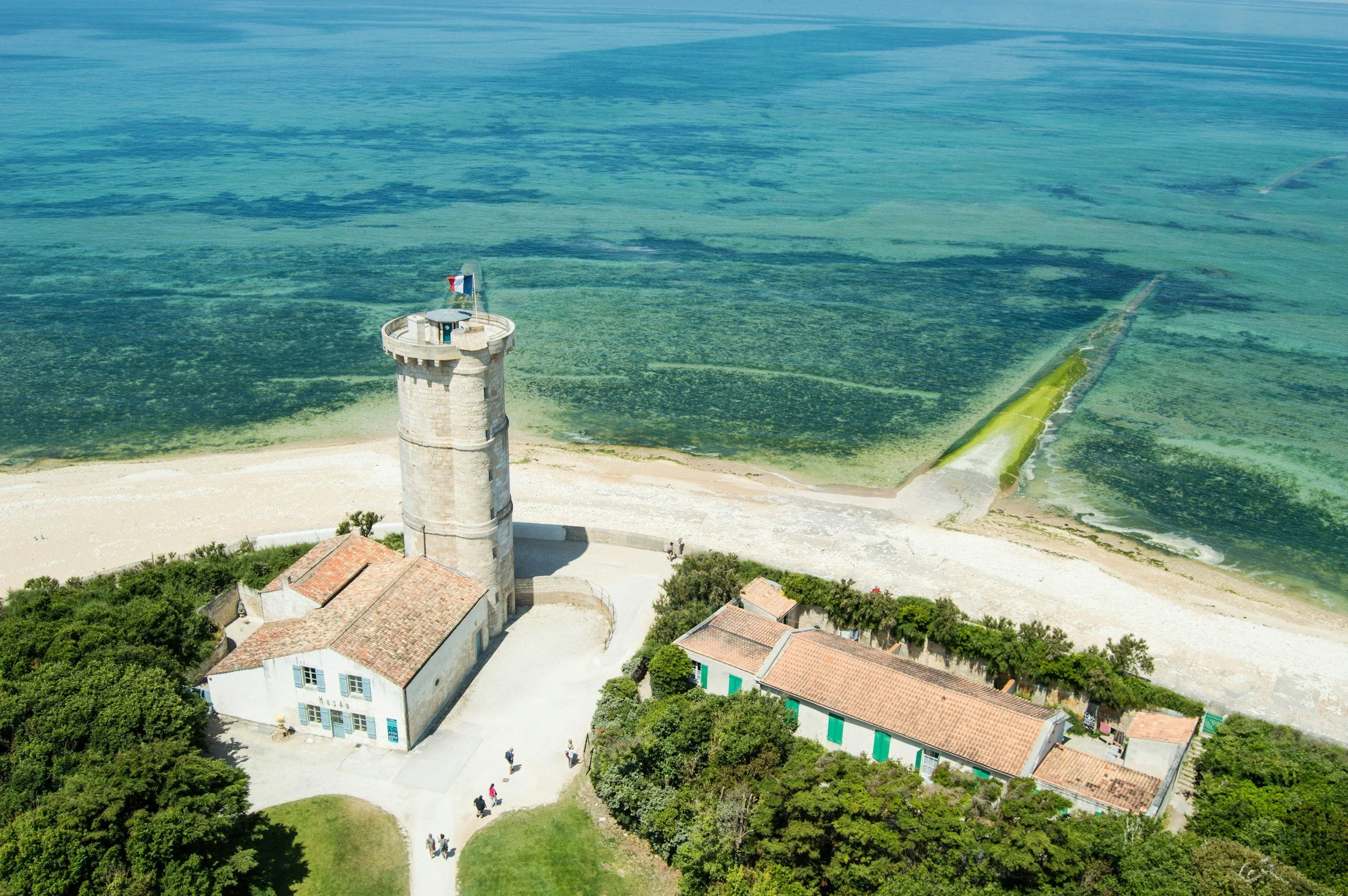 Vue aérienne d'une tour en pierre avec un drapeau français, entourée de bâtiments avec des toits en tuiles, proche de la mer avec une digue génoise à droite.