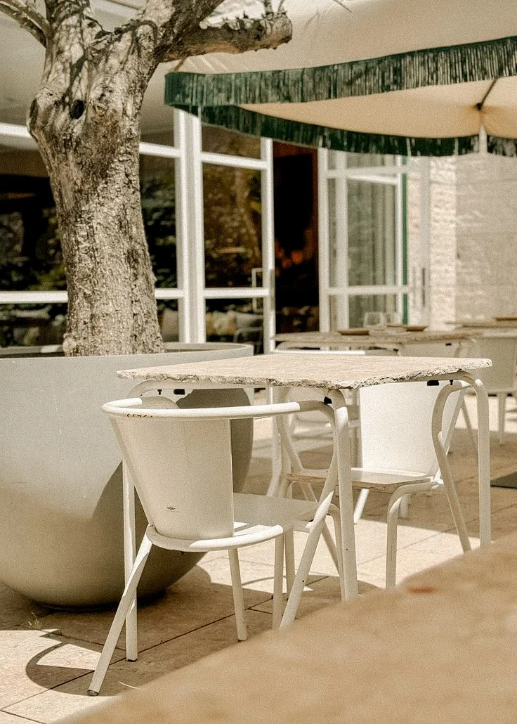 Outdoor patio with white chairs, a table, potted plant, tree, and green and beige umbrellas