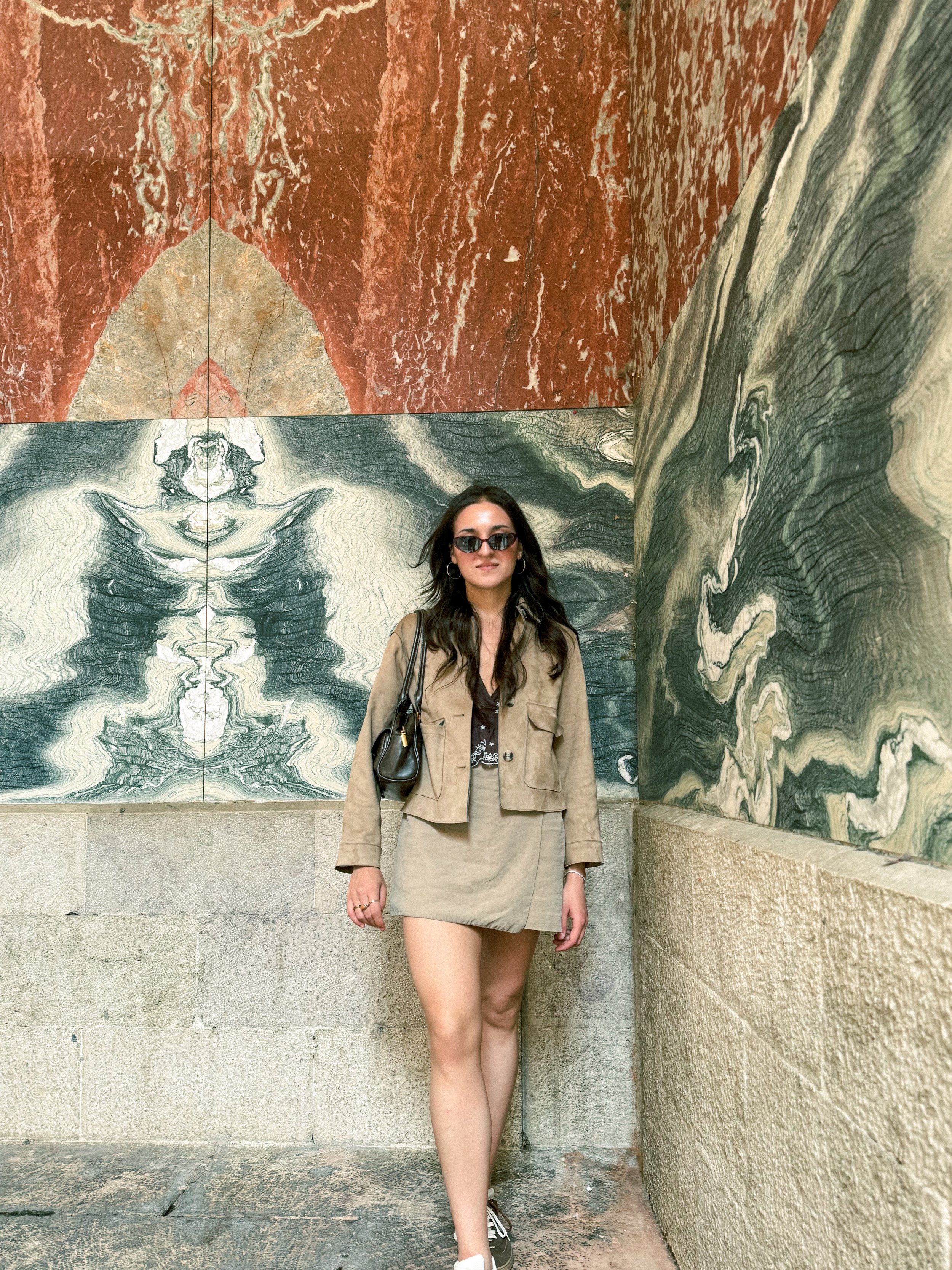 A woman with long dark hair and sunglasses standing against walls with marble-like patterns in shades of beige, gray, and rust, wearing a tan blazer, matching skirt, black top, and sneakers, with a small black handbag.