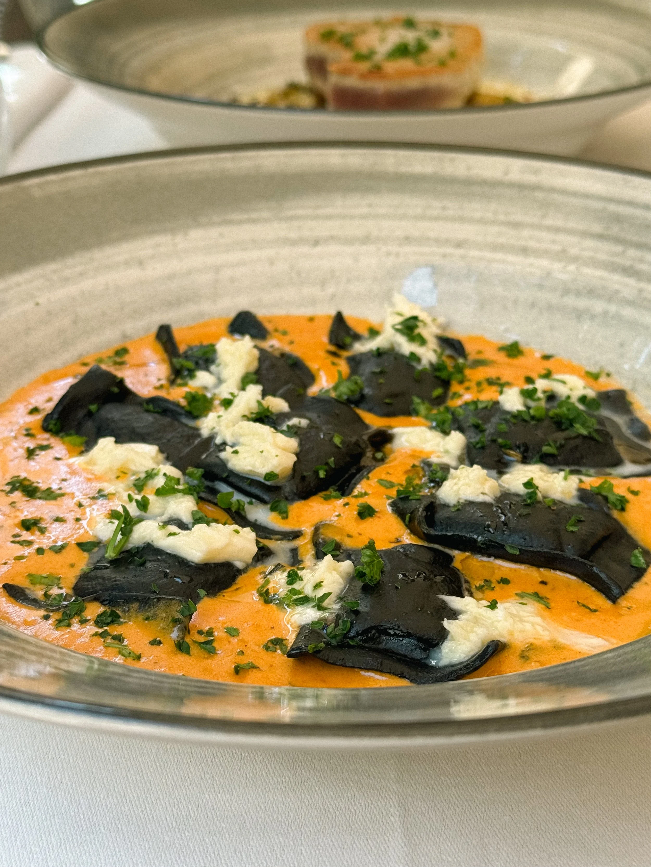 Close-up of a dish with dark mushrooms, white cheese, and green herbs in an orange sauce in a glass baking dish.