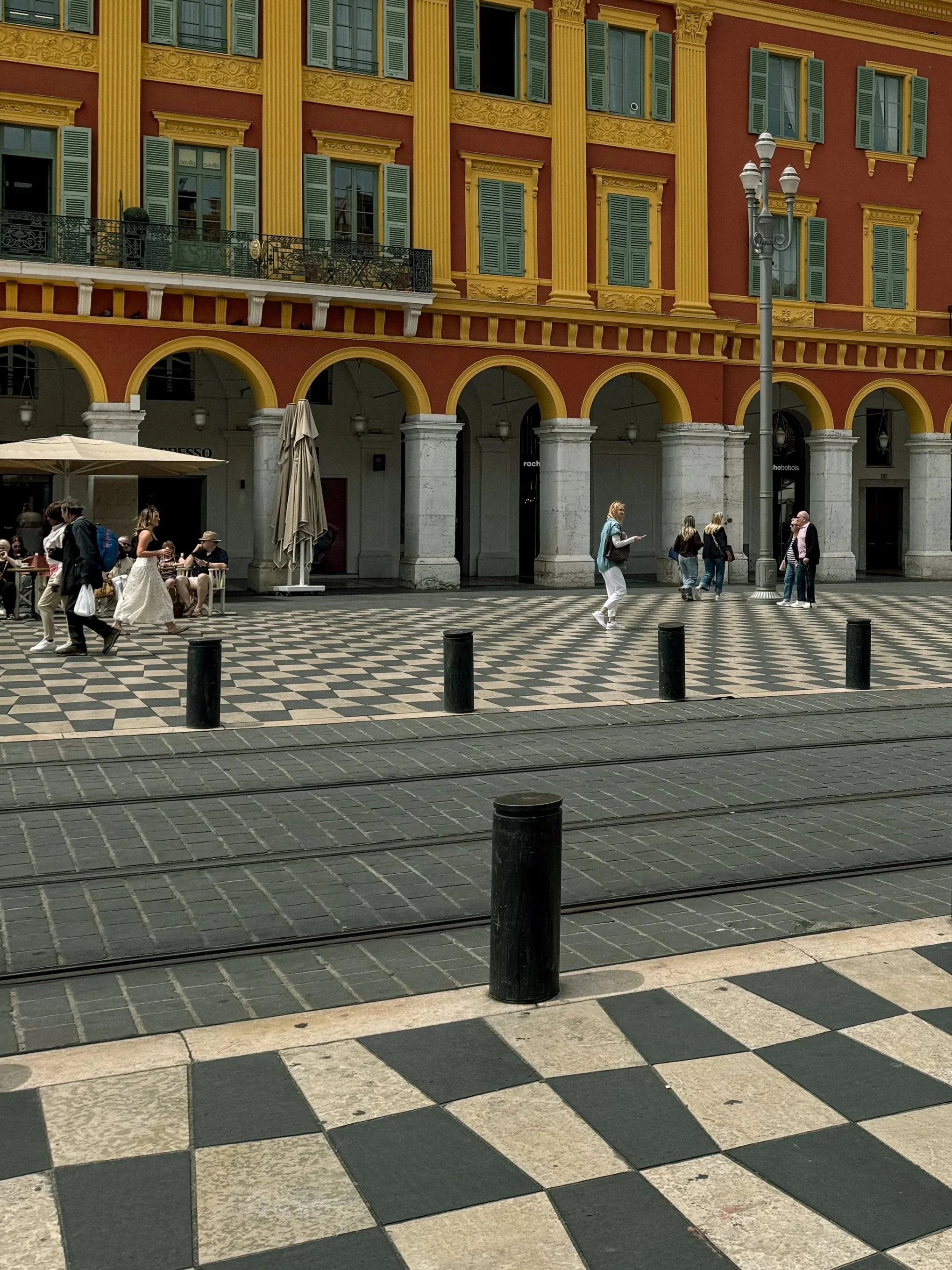 People walking and sitting outside a vibrant yellow and red historic building with green shutters, on a checkered pavement street with tram tracks and black bollards, in an urban city square.