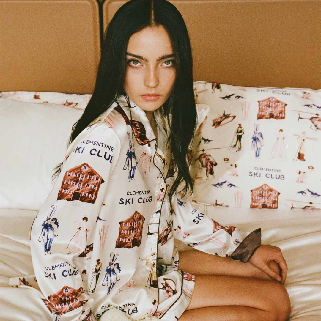 A woman with long black hair and fair skin, sitting on a bed with a pillow that has a printed design. She is wearing a cream-colored pajama set with illustrations of skiers, ski lodges, and the words "Clementine Ski Club." She has a serious expression and is looking directly at the camera.