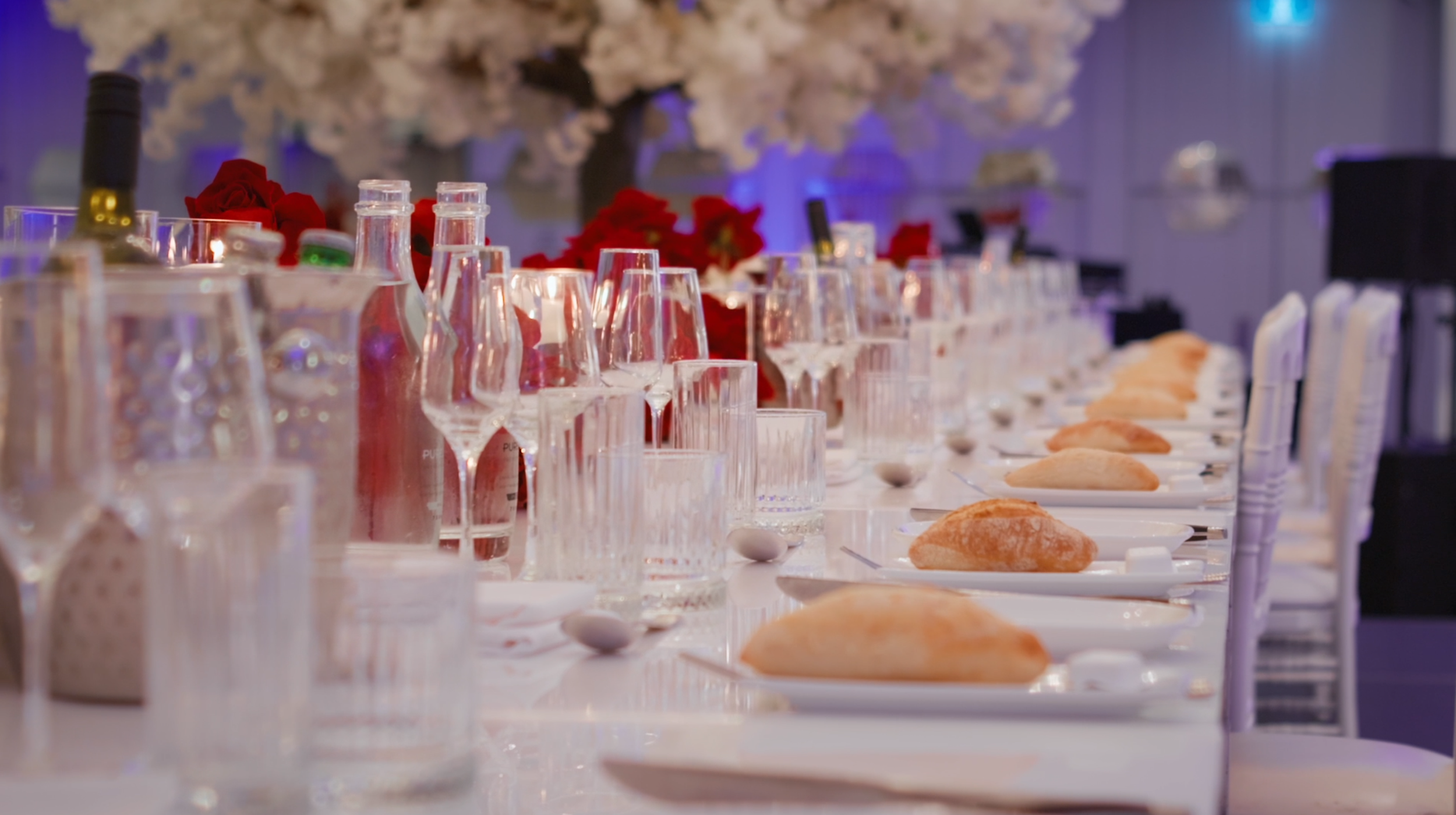Set dining table with glasses, plates, bread rolls, and decorative flowers in the background.