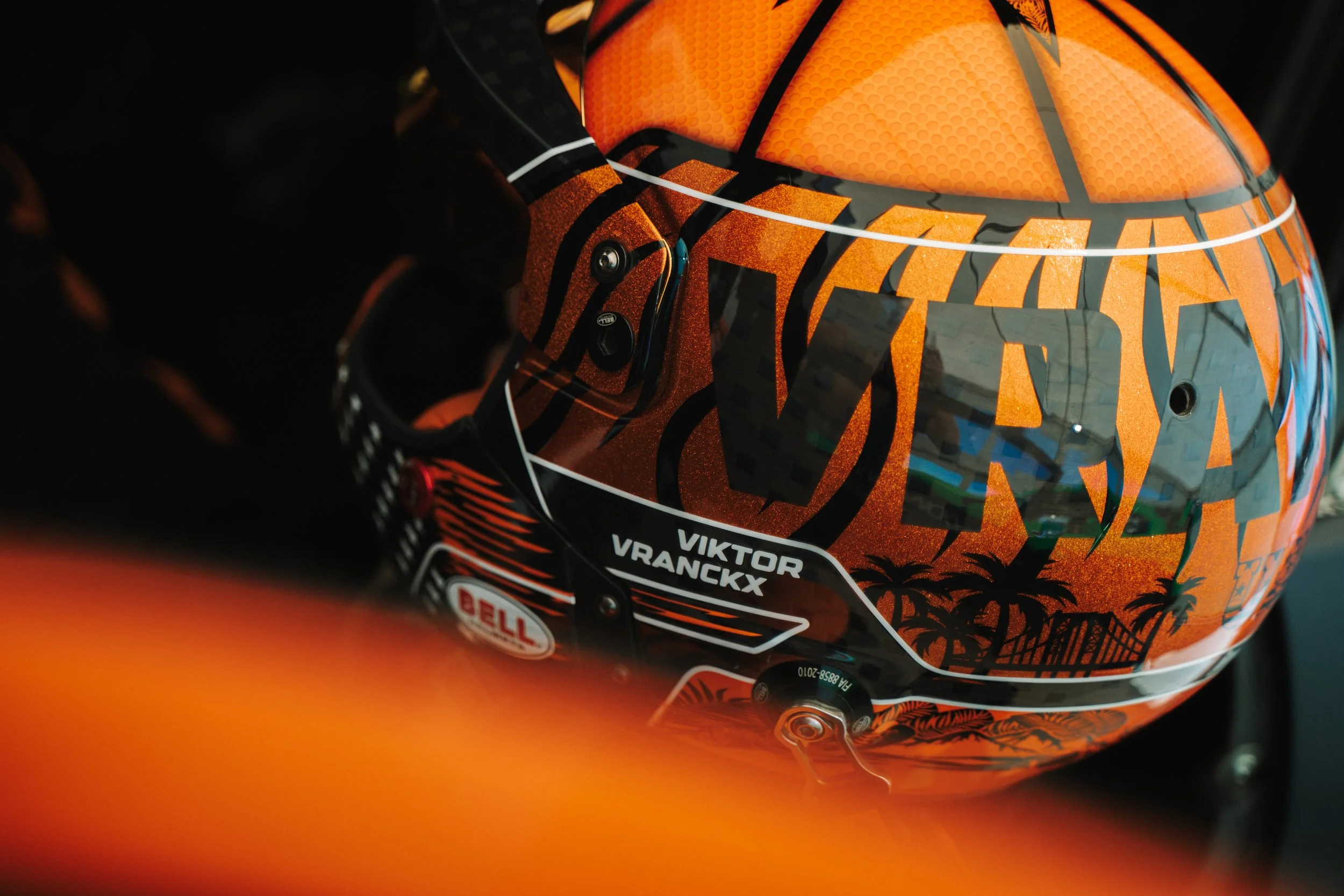 Close-up of an orange motorcycle helmet with black and orange graphics, featuring a white line design and labels including 'Viktor Vranckx' and other sponsor logos.