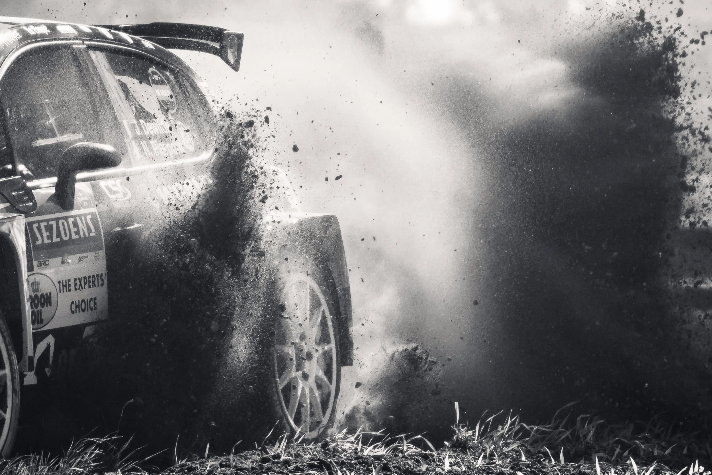 A race car speeding through a dirt track, kicking up dust and gravel, with grass in the foreground and the scene captured in black and white.