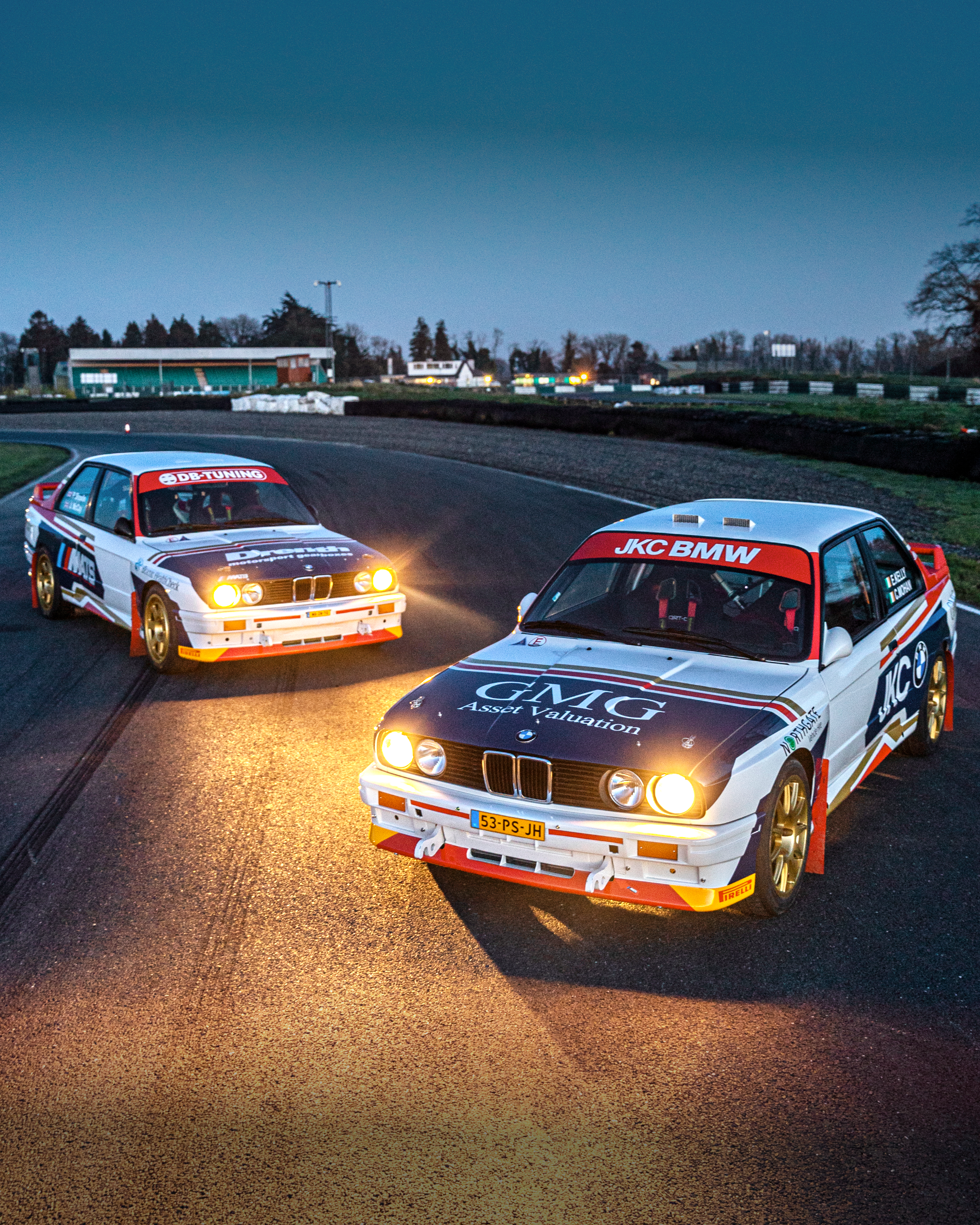 Two rally cars on a race track at dusk, with half of the cars in the foreground and a track and small buildings in the background.