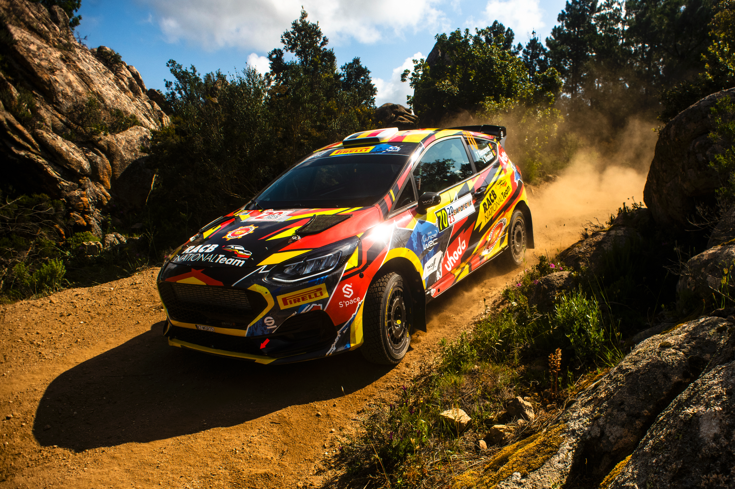 A colorful rally car driving on a dirt trail surrounded by rocks and green foliage, kicking up dust with a clear blue sky above.