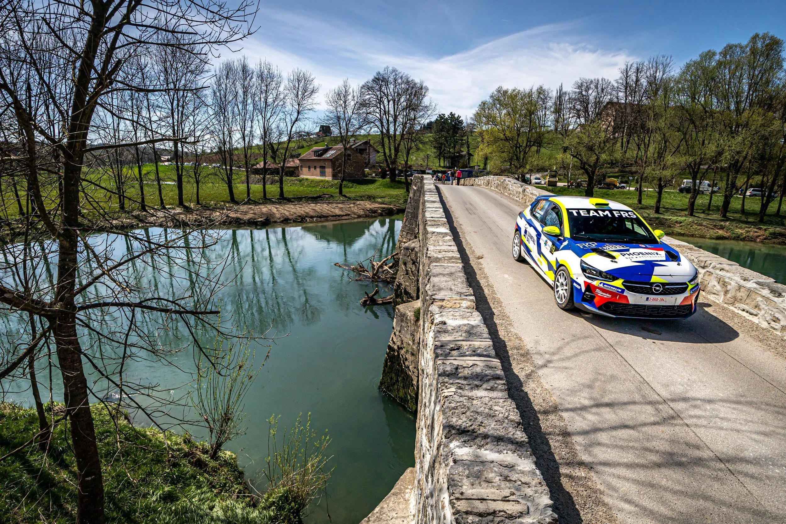 A rally car parked on a stone bridge over a river in a rural area with trees and houses in the background.