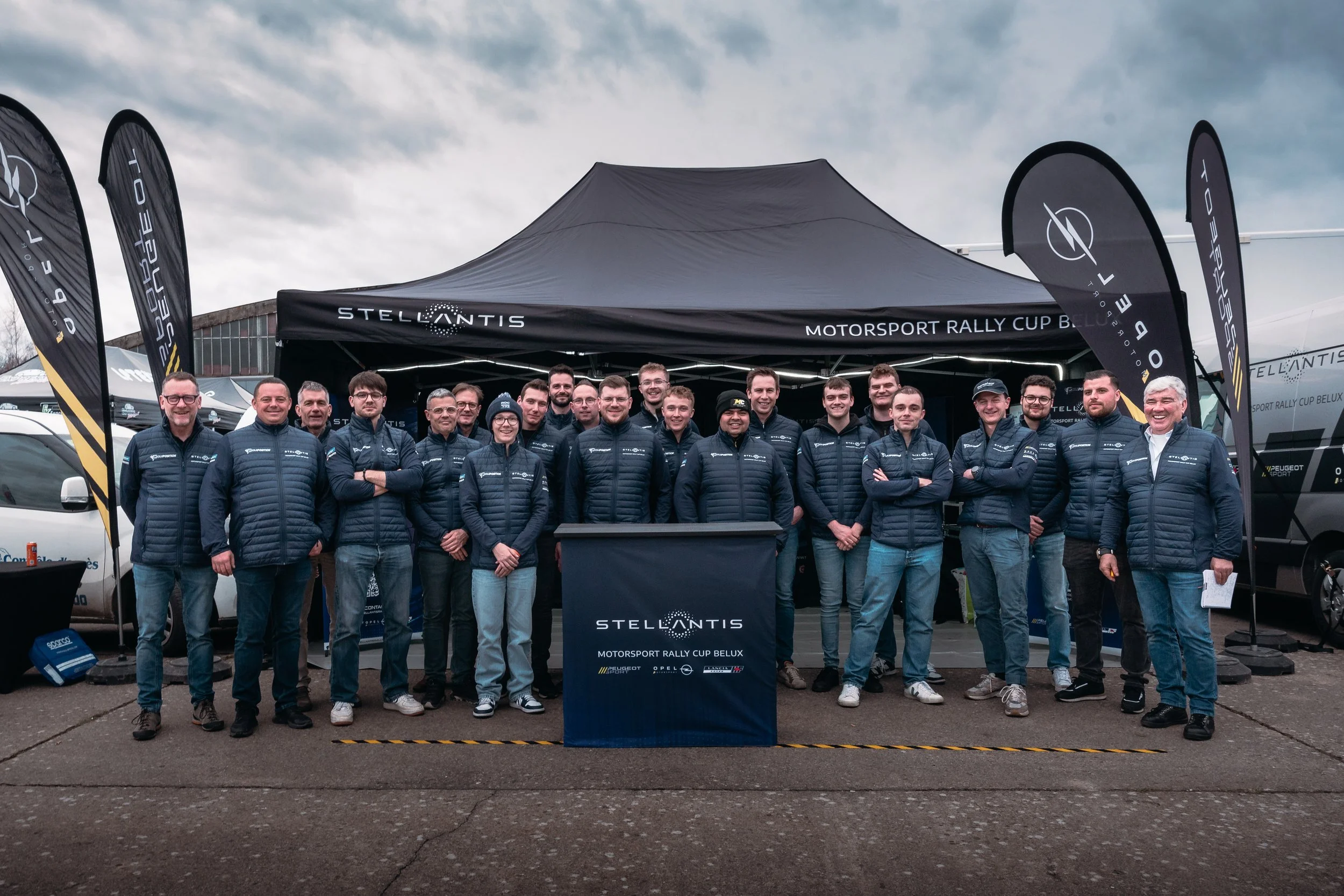 Group of people standing under a black tent that reads 'Stellantis Motorsport Rally Cup Belux'. They are wearing matching navy jackets with various logos. Flags with the Stellantis logo are on either side of the tent. The sky is cloudy.