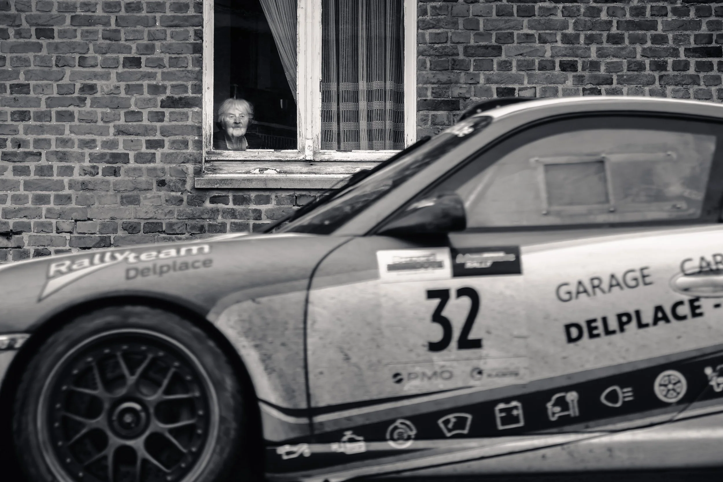 A woman looking out of a window of a brick house. In the foreground, part of a race car with various logos and the number 32 is visible.