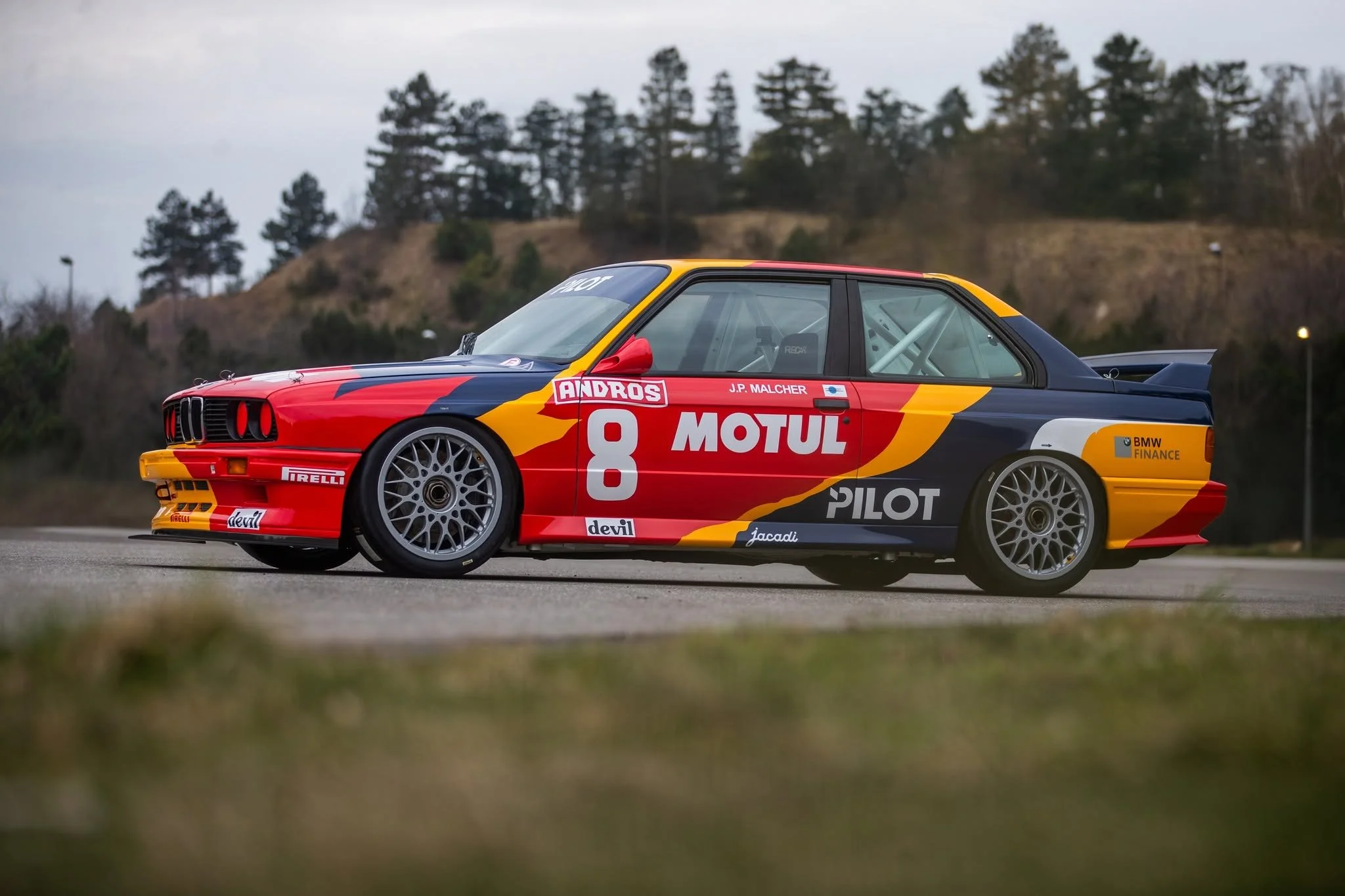 A race car with racing decals and the number 8 parked outdoors on a paved surface, with a background of trees and a cloudy sky.
