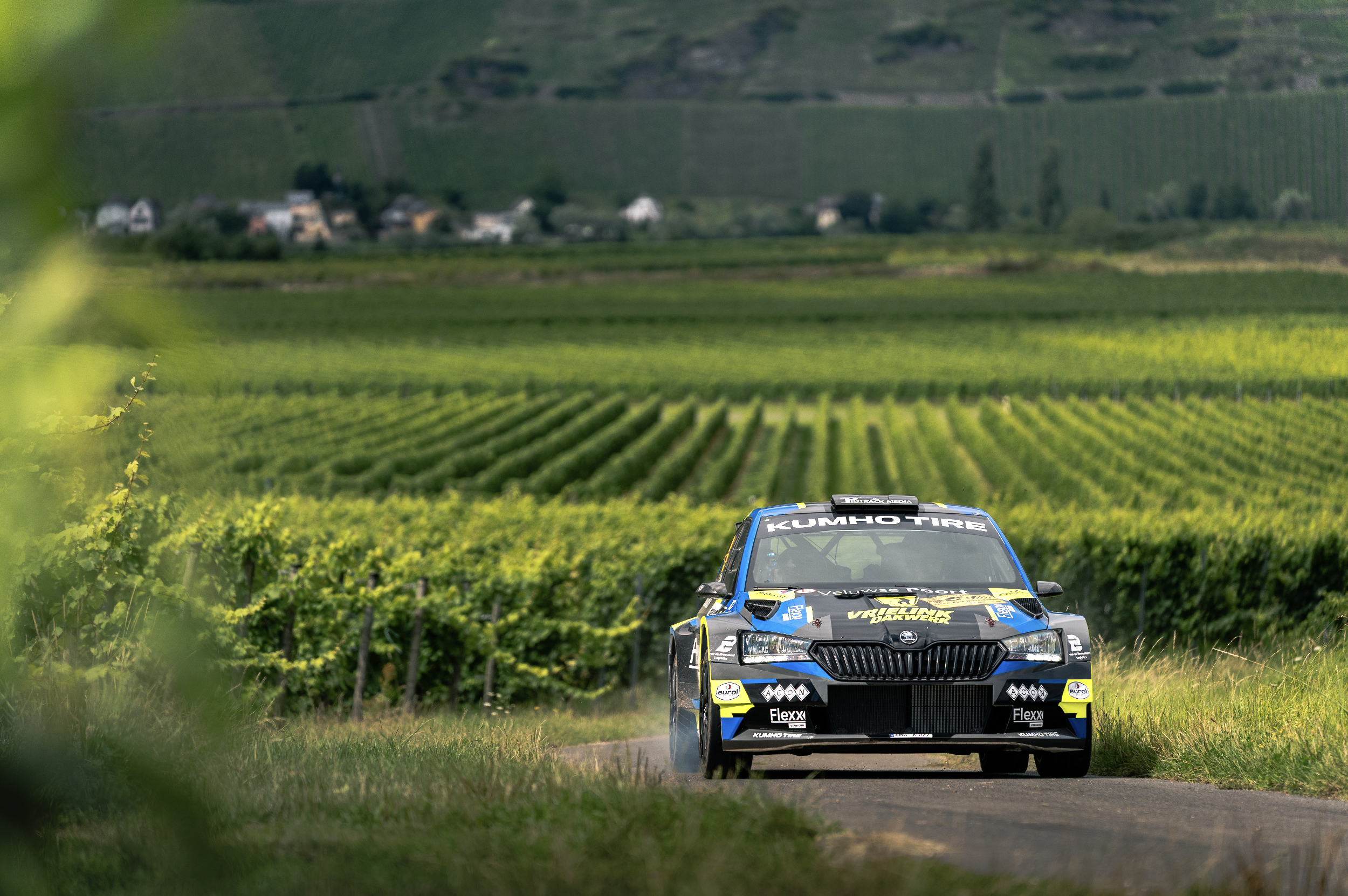A rally race car driving along a rural road surrounded by green vineyards with hills and a small village in the background.