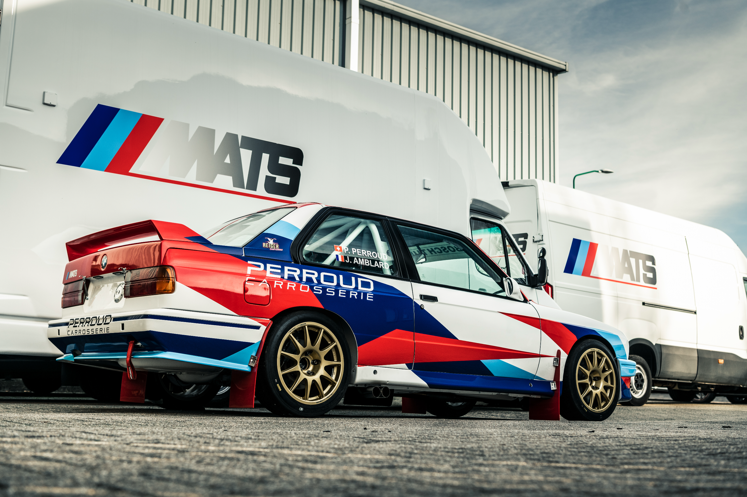 A racing car with a red, white, and blue livery featuring the names P. Perroud and J. Amblard on the window, parked in front of White trucks with the LMS logo, and located on a paved surface outdoors with a cloudy sky in the background.