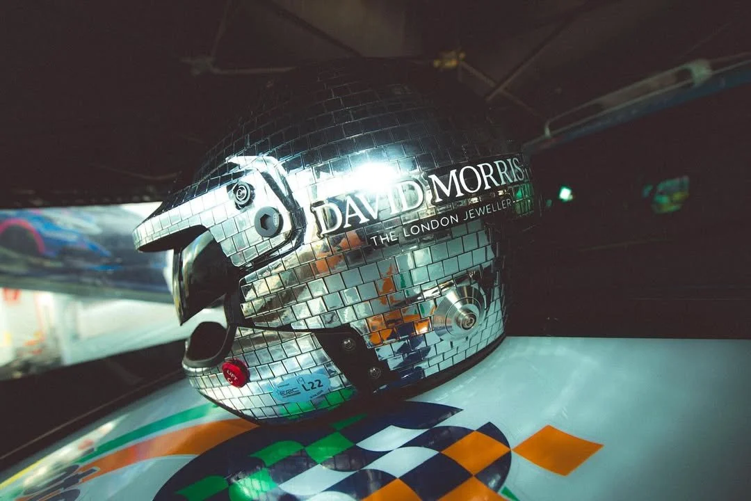 A shiny, mirror-like motorcycle helmet with a mosaic pattern, featuring the text 'DAVID MORRIS THE LONDON JEWELLER,' placed on a colorful surface inside a vehicle.