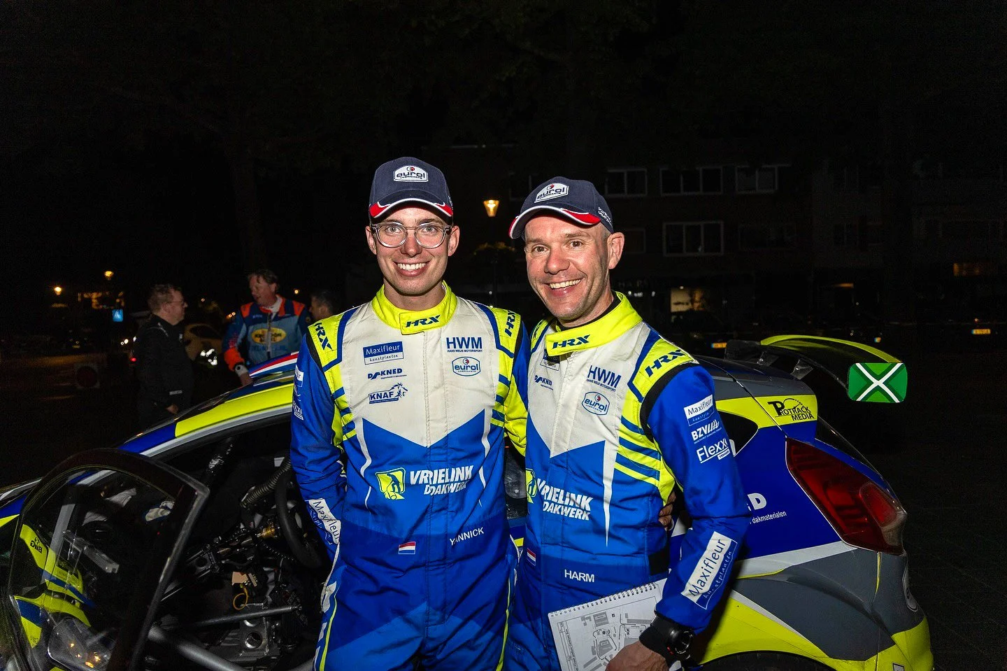 Two rally drivers in blue and yellow racing suits standing in front of a rally car at night, smiling for the camera.